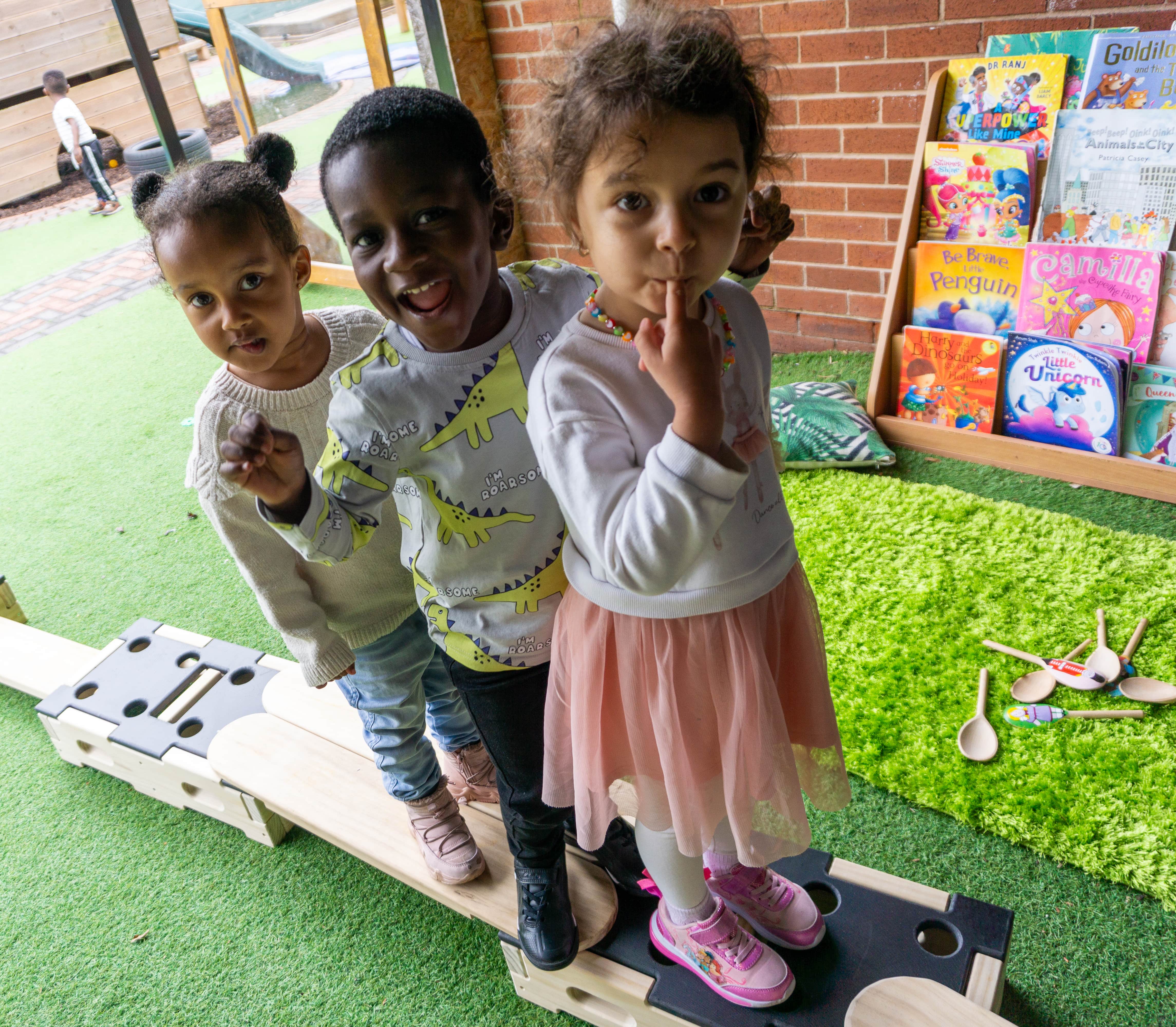 3 children are stood in a single file line as they all look at the camera. They are stood on wooden Accoya planks and boxes as they all pose for the camera. The Accoya wooden equipment is set up on artificial grass.