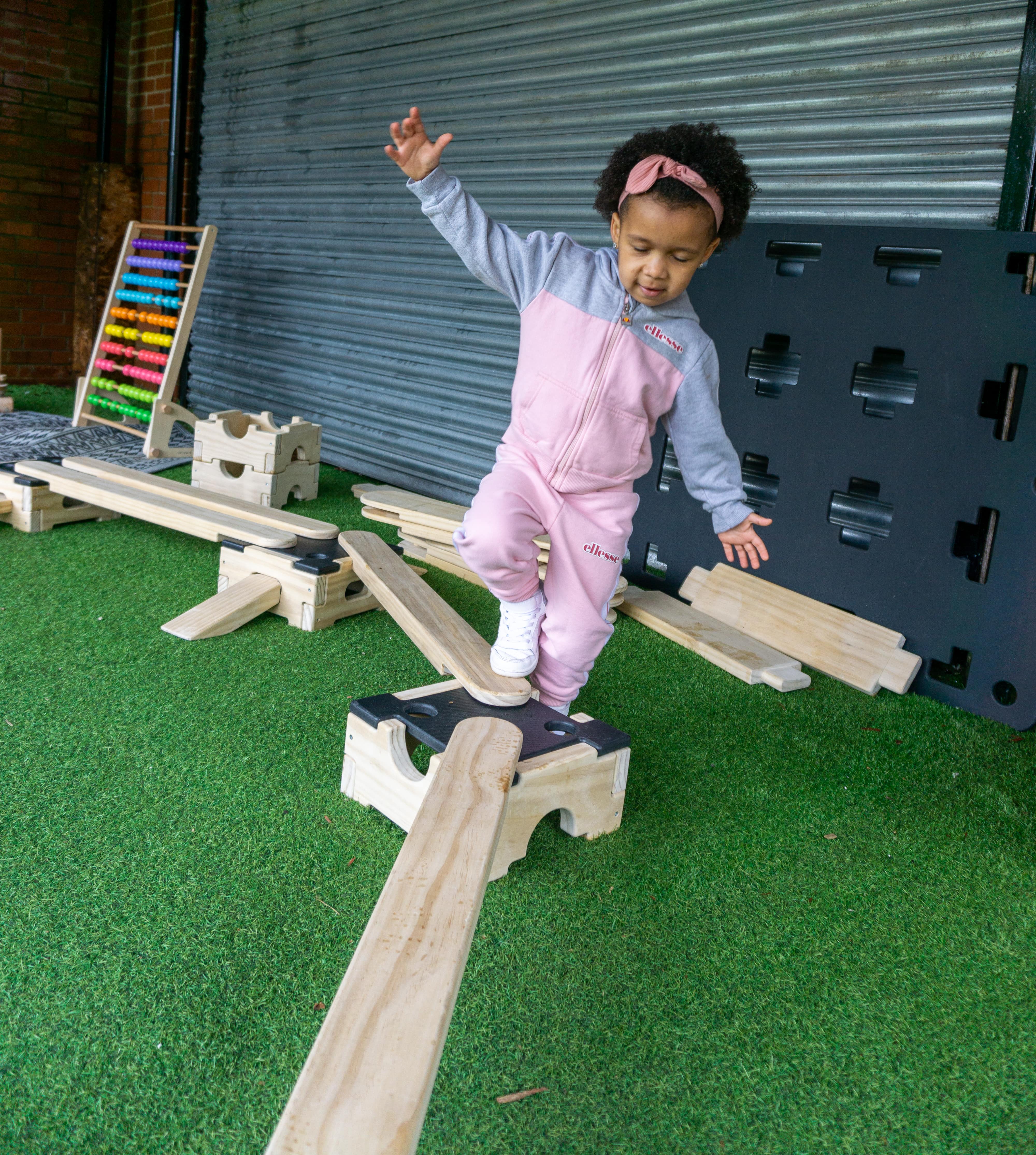 A little girl is crossing an Accoya wooden plank as she tries to make it to an Accoya wood block. The small obstacle course is set up on artificial grass.