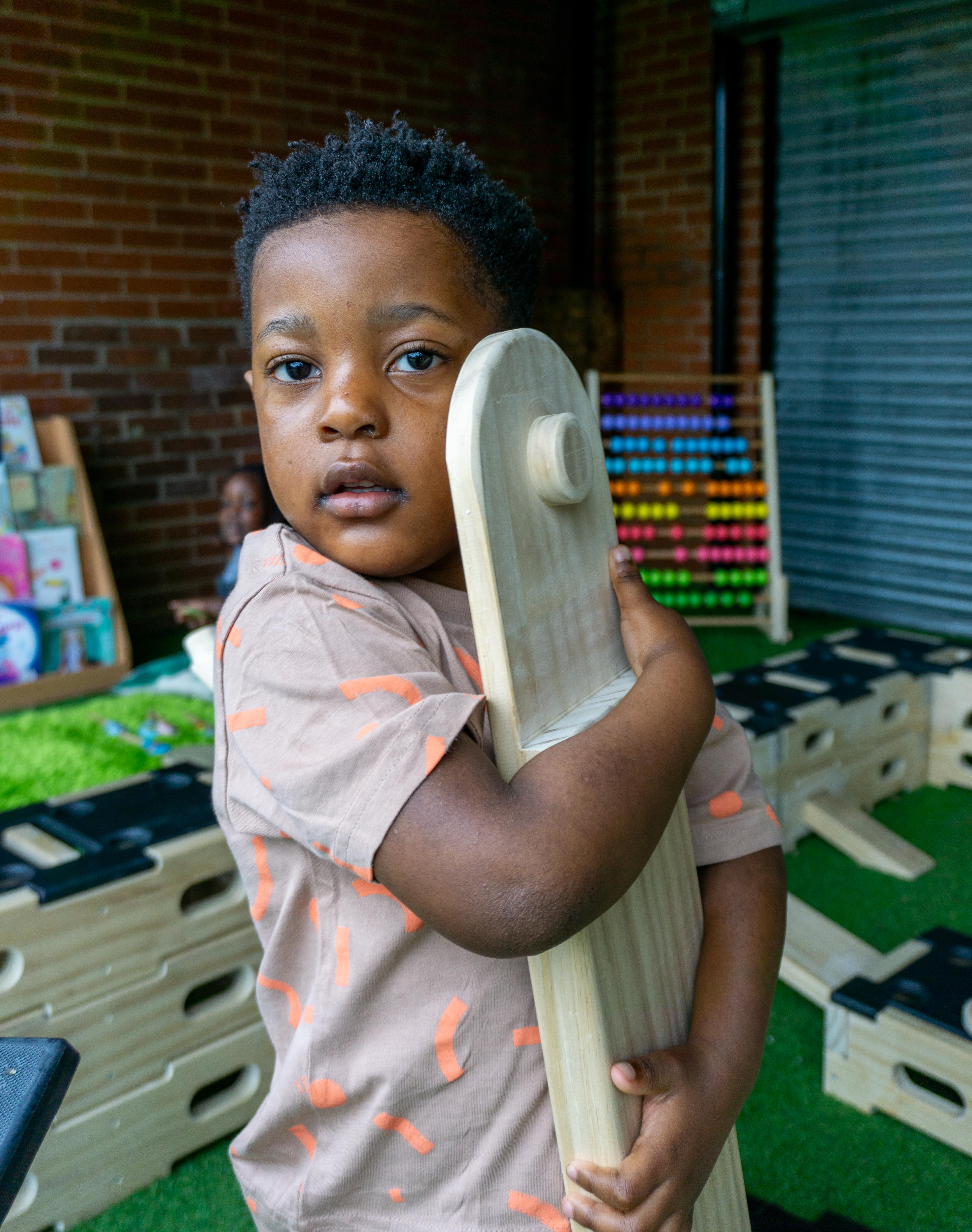 A little boy hugging a plank of Accoya wood, holding it close to his cheek. The boy is looking at the camera as other Accoya-based wooden equipment can be seen in the background, all on top of an artificial grass surfacing.