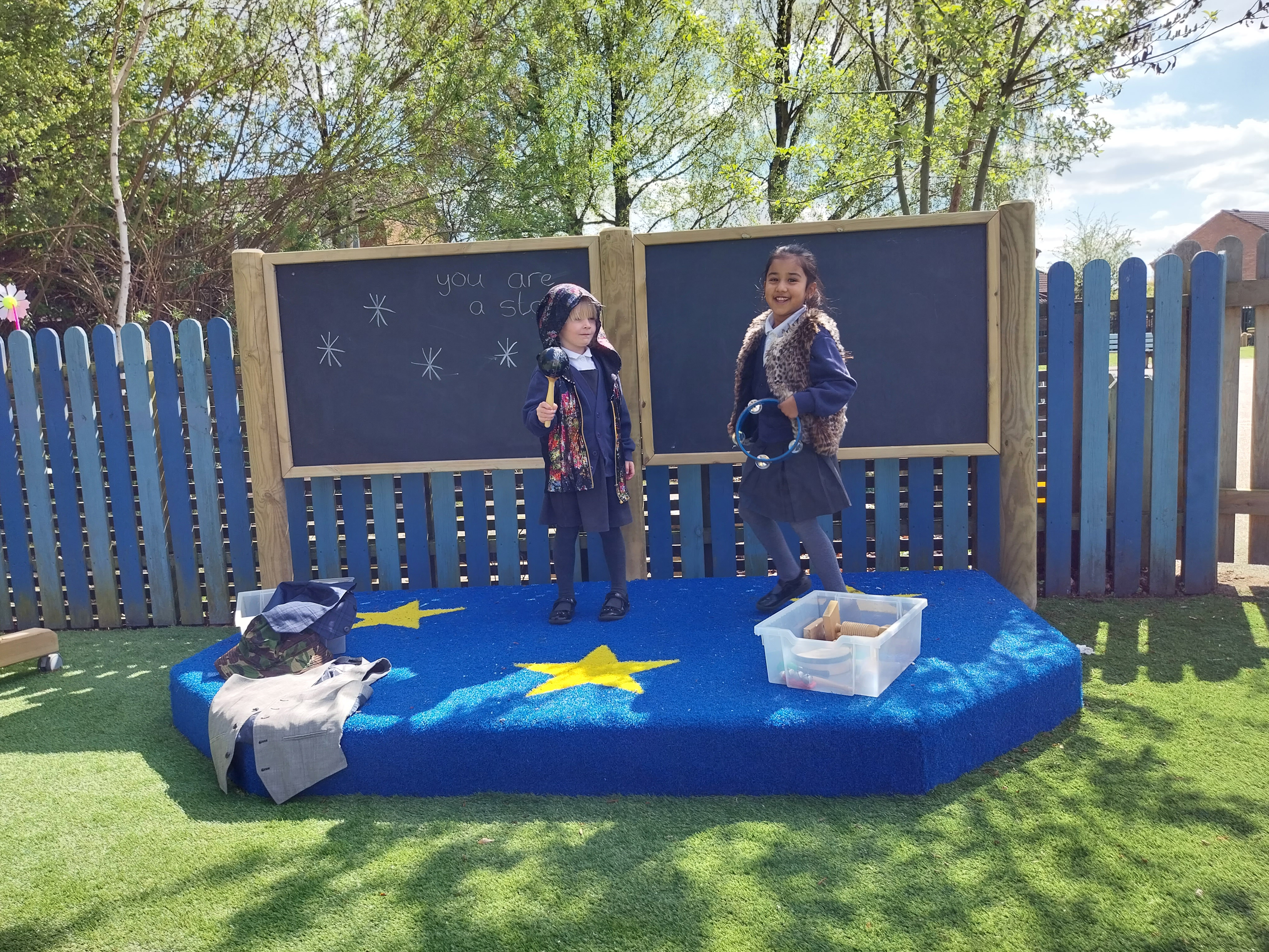 2 children are stood on a small stage that has three wooden posts placed behind it, with a chalkboard placed between each one. The stage is a semi-circle with a blue saferturf surfacing. There are yellow Saferturf stars placed on the stage too. The two children are looking at the camera as they hold props and the chalkboard shows "you are a star" on it.