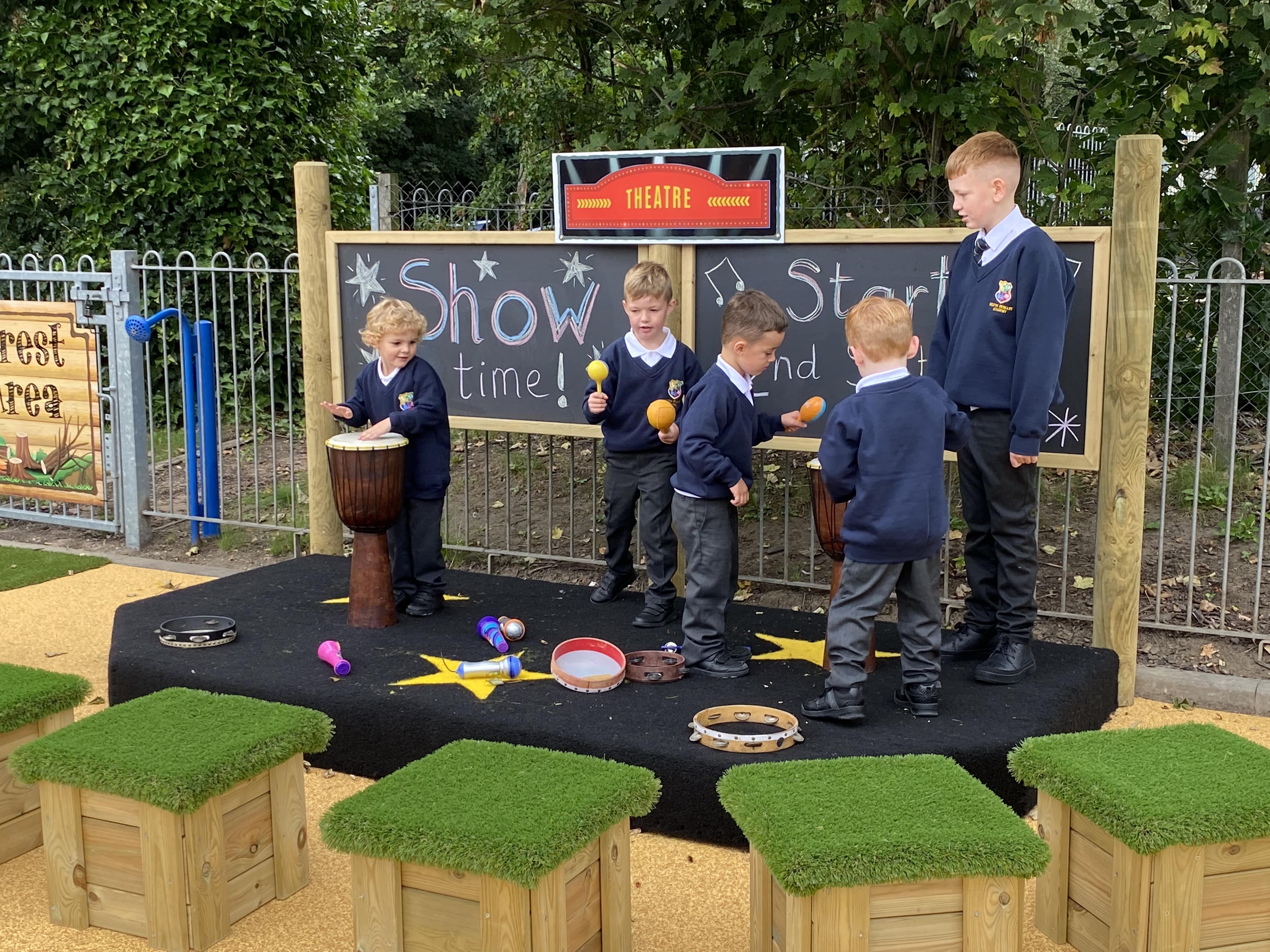 5 children are stood on a performance stage, which is covered in black Saferturf. They're holding a variety of instruments as they perform on the stage. On the chalkboard placed behind them is the words "Show time" and a sign that says "Theatre". 5 wooden blocks are placed in front of them, with artificial grass tops. They are designed to be seats.