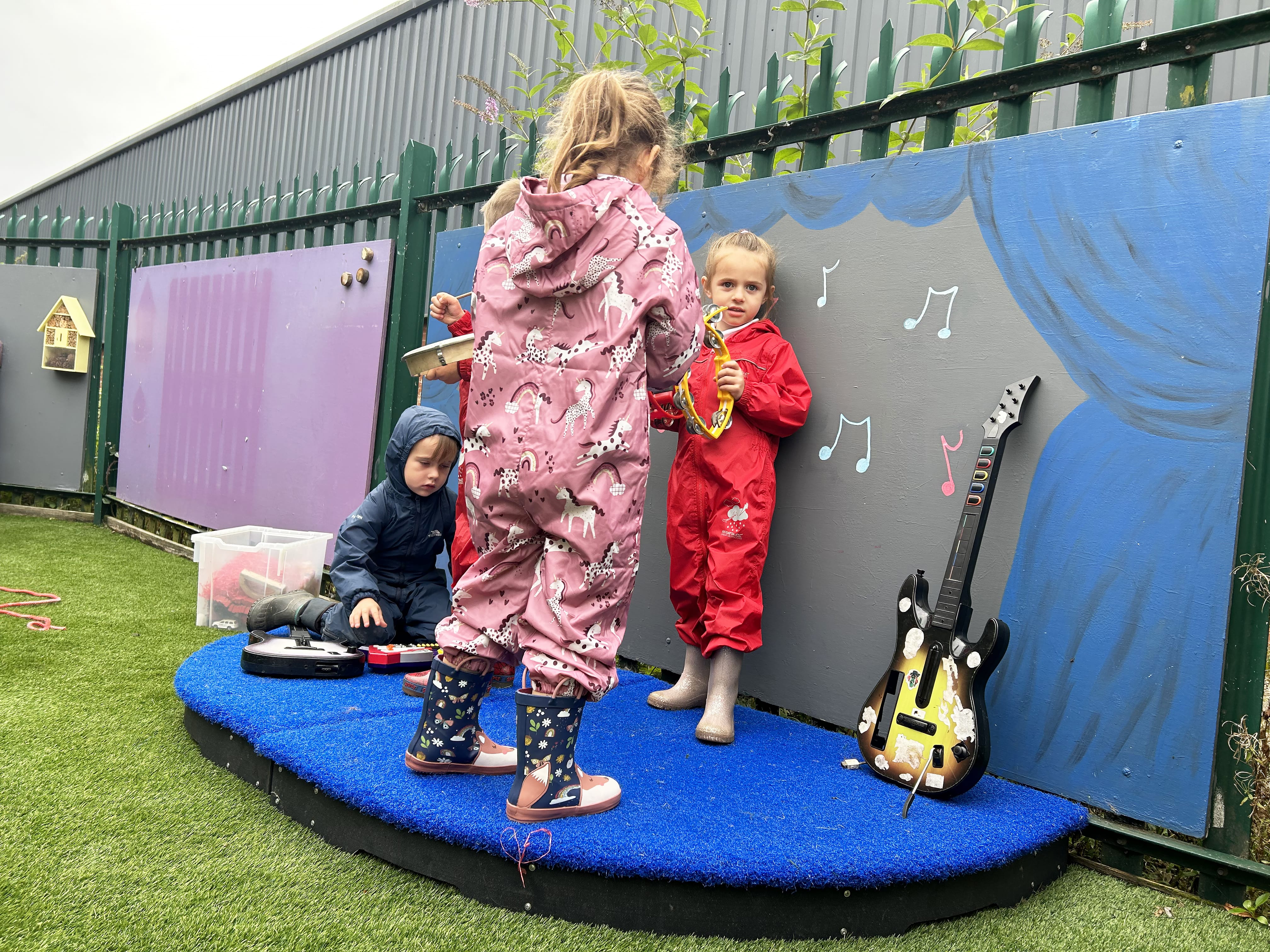 4 children are stood on a small, semi-circle stage as they interact with a variety of musical instruments. The background of the stage is a painting of theatre curtains with musical notes in the middle.