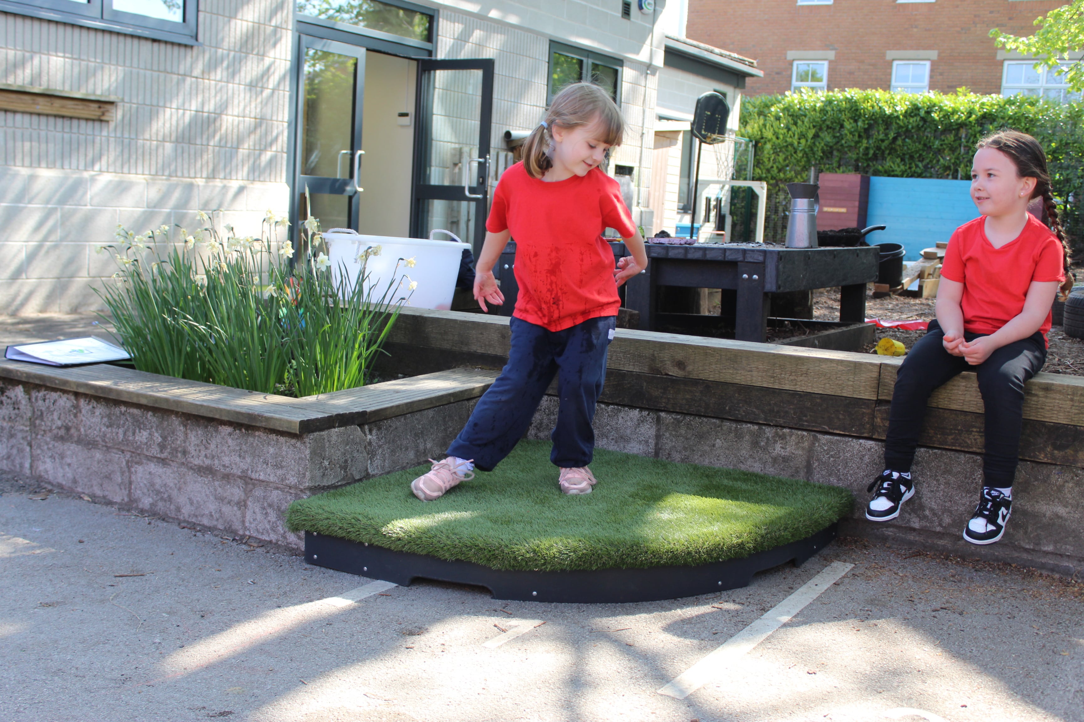 A small grass stage is placed in a corner outside. The stage has black sideboards but an green artificial grass surface. A little girl is stood on top of the stage and is dance. Another little girl can be seen sitting on the wall next to her and watching. 