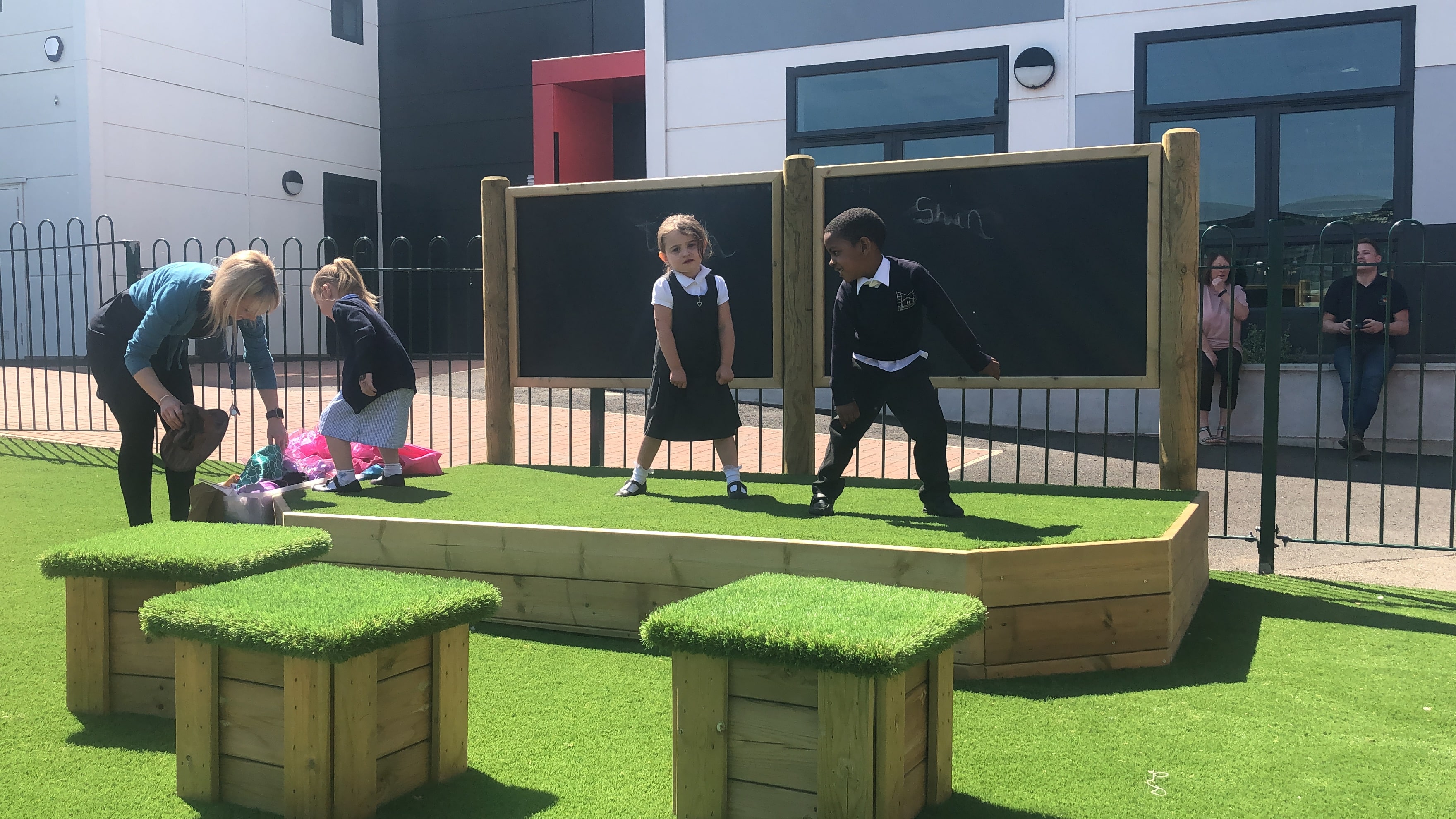 Two children are dancing on the outdoor stage, with a third child stood to the right of the stage and is looking for a prop or outfit to use. The stage has an artificial grass surface to match the artificial grass surfacing on the playground and the wooden blocks with artificial grass placed on top of them.