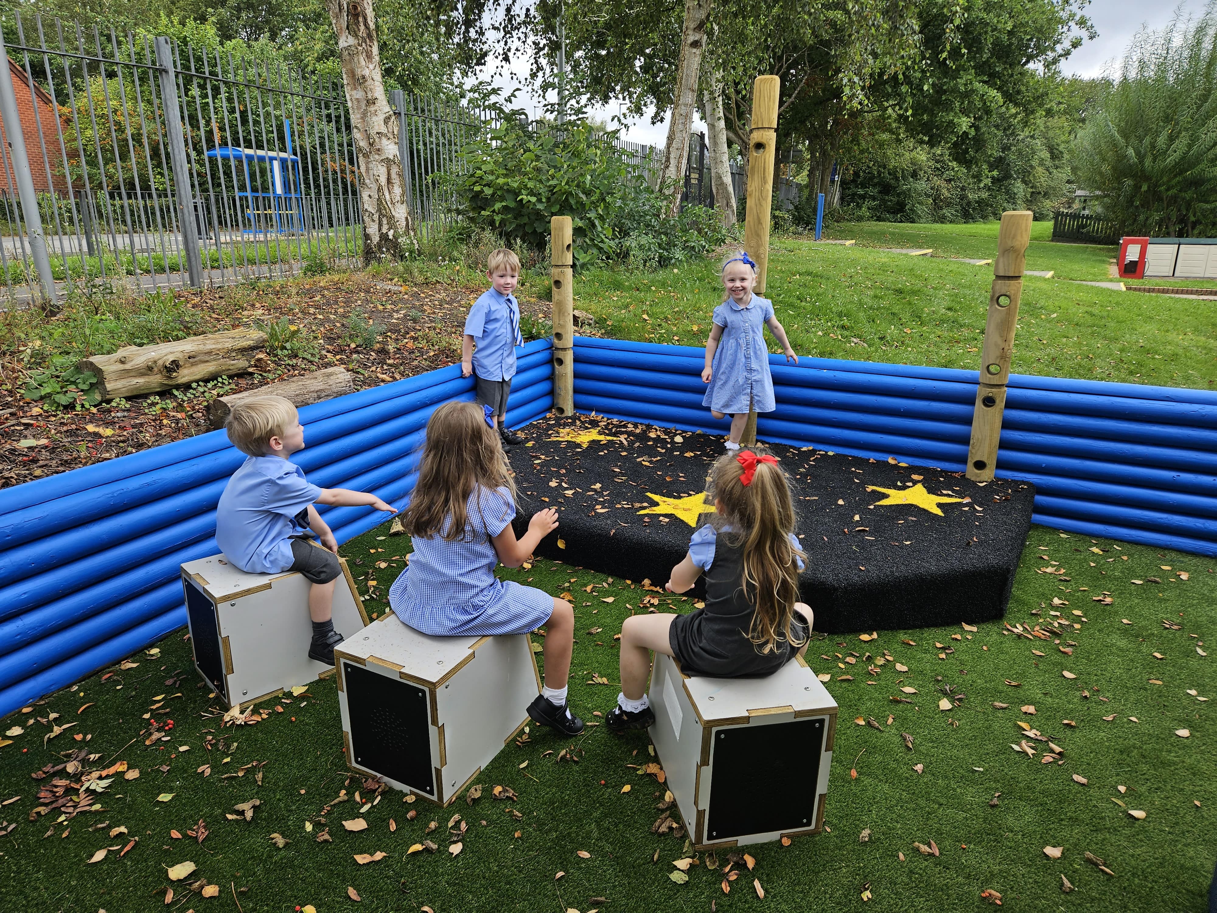 2 children are dancing on a black stage with yellow stars placed on it. 3 children are sat on drum seats and are playing a song as the children rehearse.