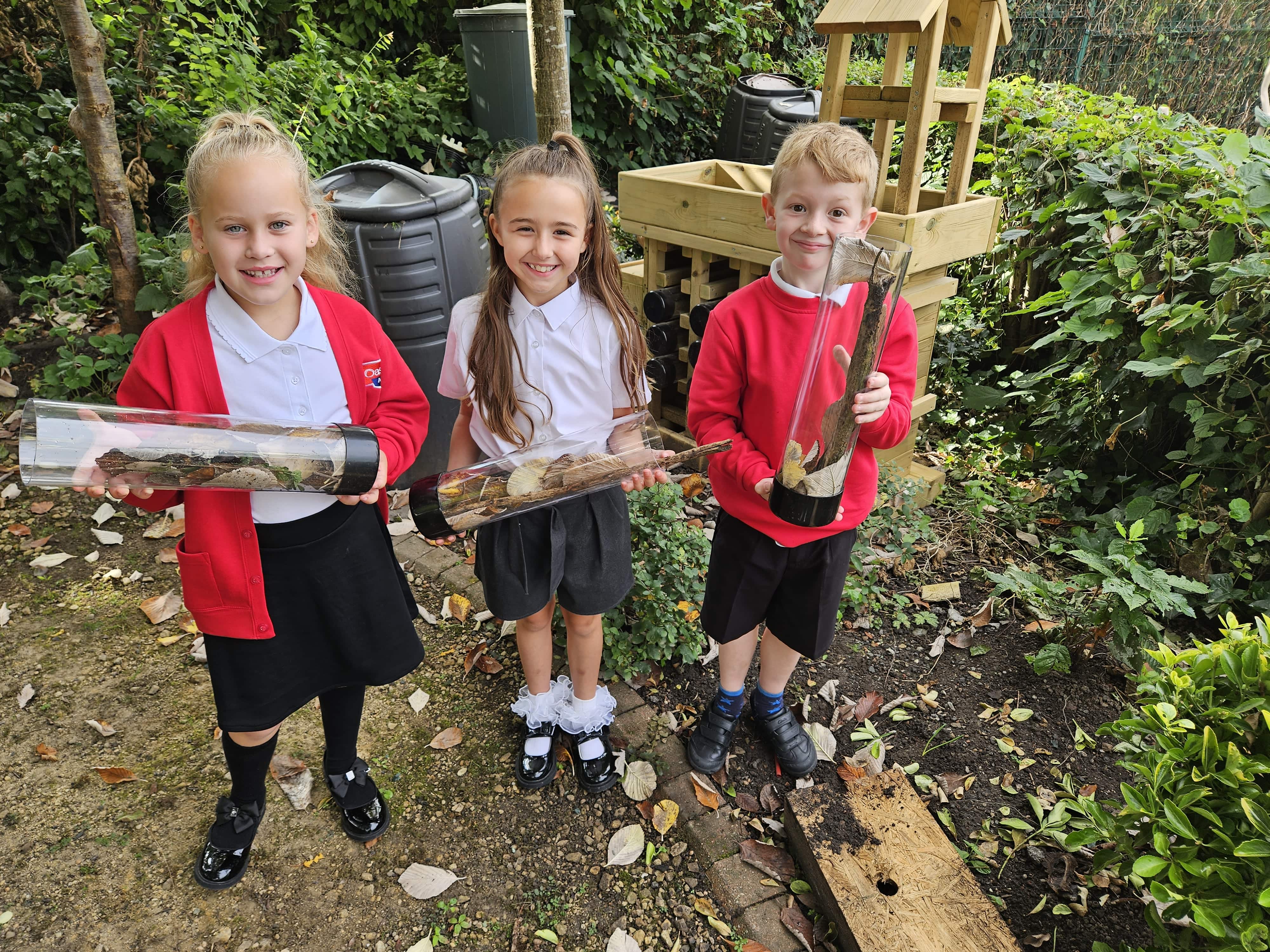 Three children are facing the camera as they hold plastic tubes which contain natural materials like sticks, leaves and mud. The children look excited as they stand in front of bushes and foliage. 