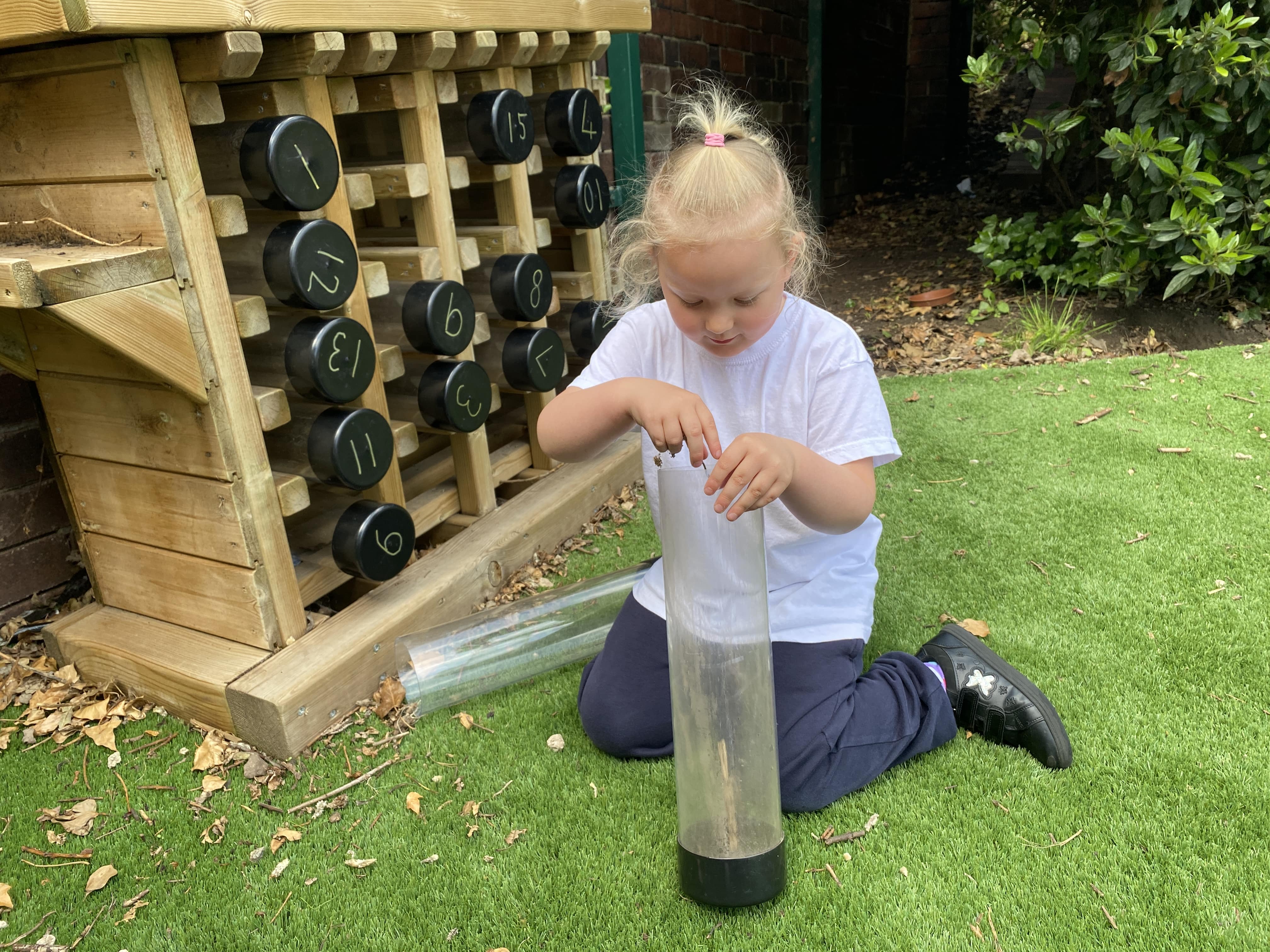 A girl is on her knees as she fills up a plastic tube with natural materials. Behind her is a wooden structure which holds these plastic tubes, called the Bug Hotel. Each tube has a number written on it, allowing for identification to be easy.