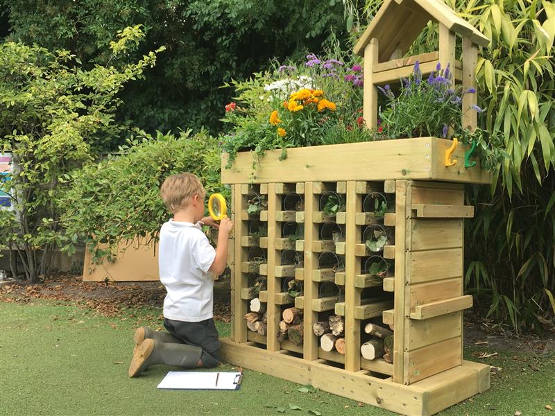 A child is on their knees as they look through a magnifying glass. The child is looking into gaps within a big, wooden structure. This wooden structure is called the Bug Hotel and is placed around bushes and flowers.