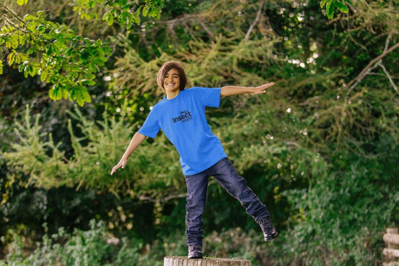 A woman is stood on top of a wooden stump, balancing on one leg. She has her arms out wide and the other leg lifted on an angle. She is wearing a blue top which says "Insect Week" on it. The background is a variety of trees and plants.