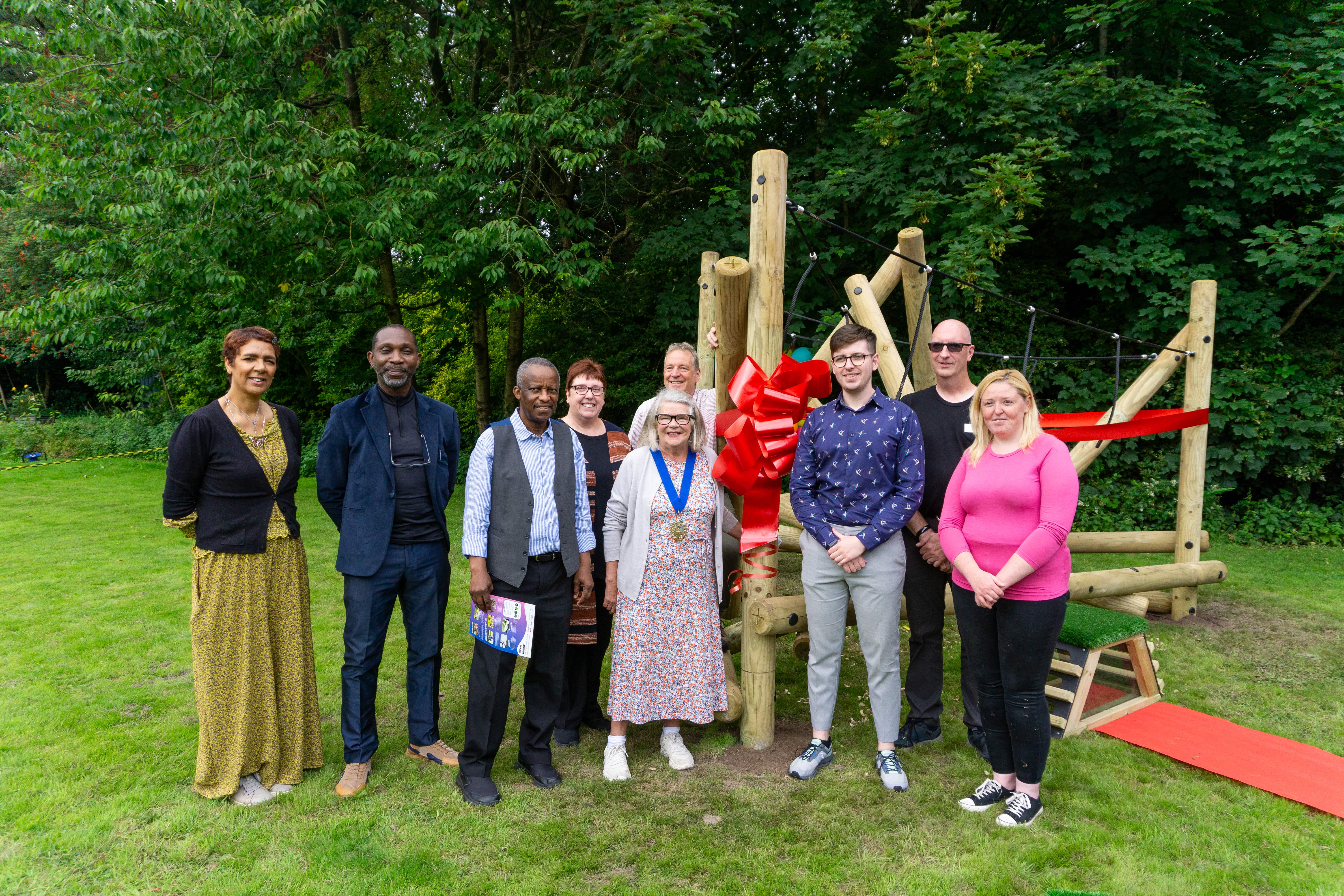 A group of 9 people are stood in front of the Latrigg Climber, a piece of wooden play equipment, that has been donated to the Bright Park by Pentagon Play. It is surrounded by trees and grass.