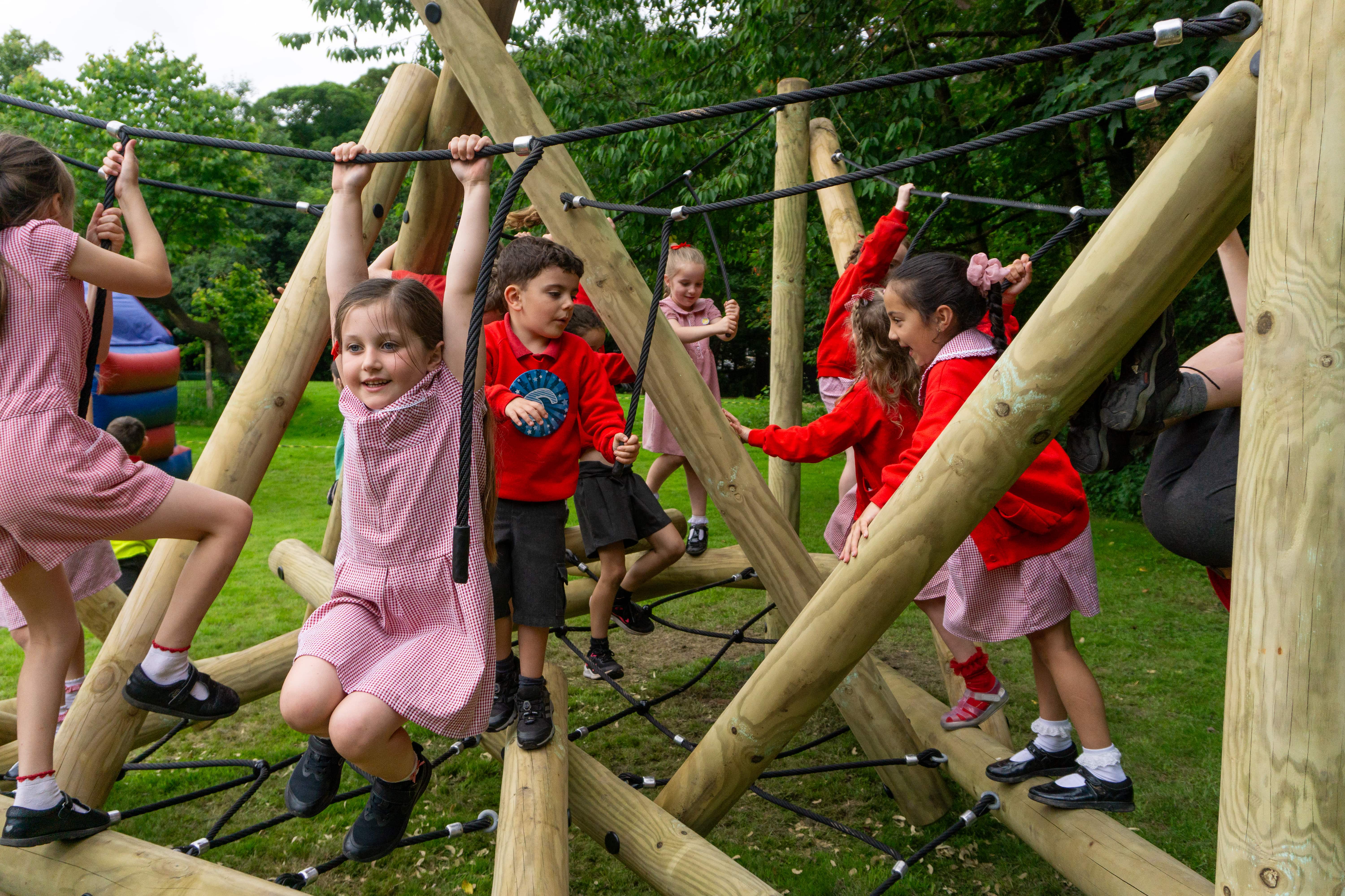 A group of children are playing on the Latrigg Climber, a wooden climbing frame that has been designed by Pentagon Play. The children are excited and happy as they play on it.