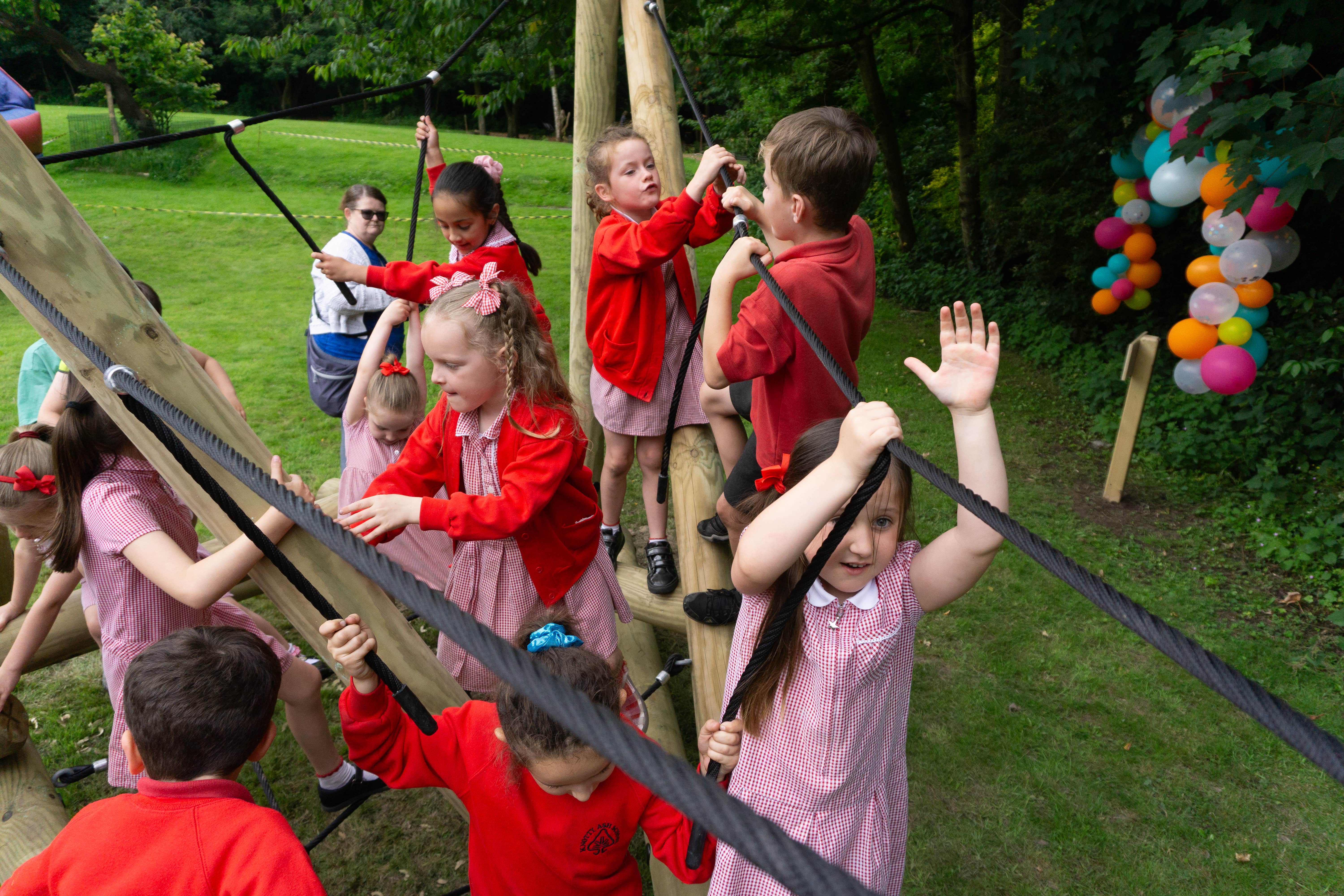 A group of children are playing on a wooden climbing frame, with ropes connecting the logs together. The children are smiling and laughing as they climb across the structure.