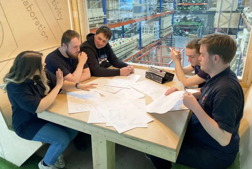 5 of our product designers sat together during a design meeting and talking through different ideas. They have a lot of designs laid out on the table as they talk to each other.