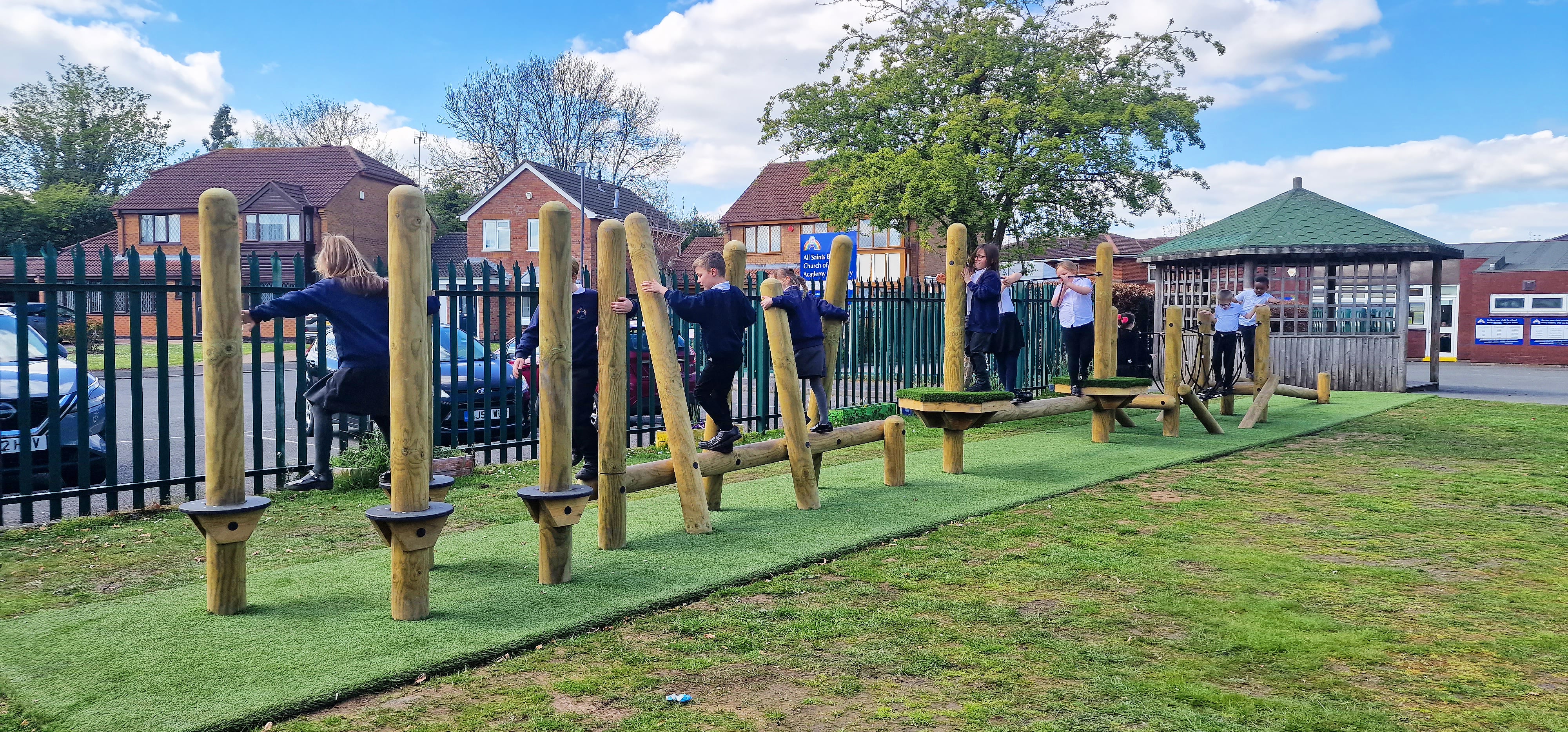 A trim trail that has been installed on an artificial grass surface, which is on top of a natural grass surface. A group of children are playing on the equipment and are making their way through the course in a single file line.