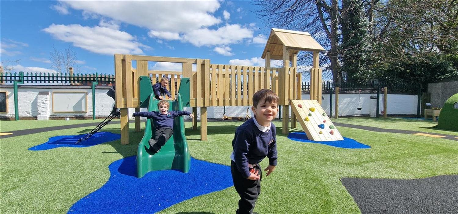 Three children are playing on a playtower that has been installed on a artificial grass surface. Some blue patches of Saferturf have been installed underneath the slide, the rope ladder and climbing wall. A black wetpour path is going around the play area.