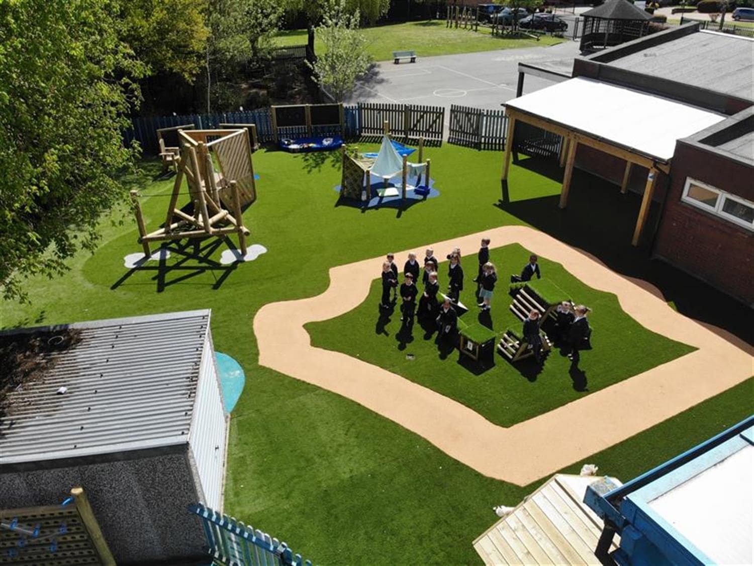 A birds-eye shot showing an artificial grass playground with a wetpour path that has been installed on top. A Playframe, den making posts, a performance stage and other play equipment can be seen across the playground.