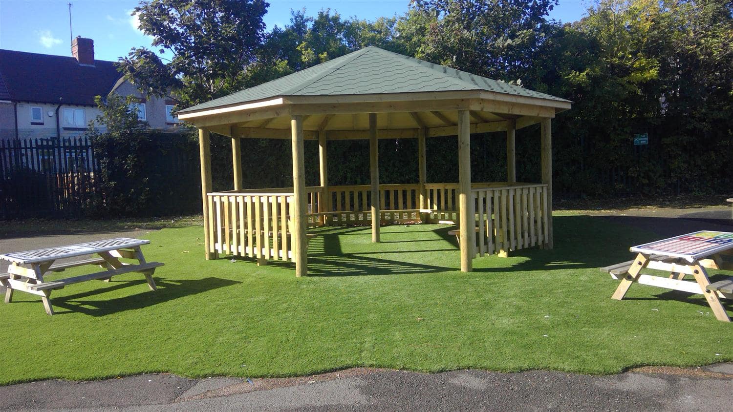 A wooden gazebo that has been built in the middle of an artificial grass play area. Two picnic benches can be seen on either side of the wooden structure.