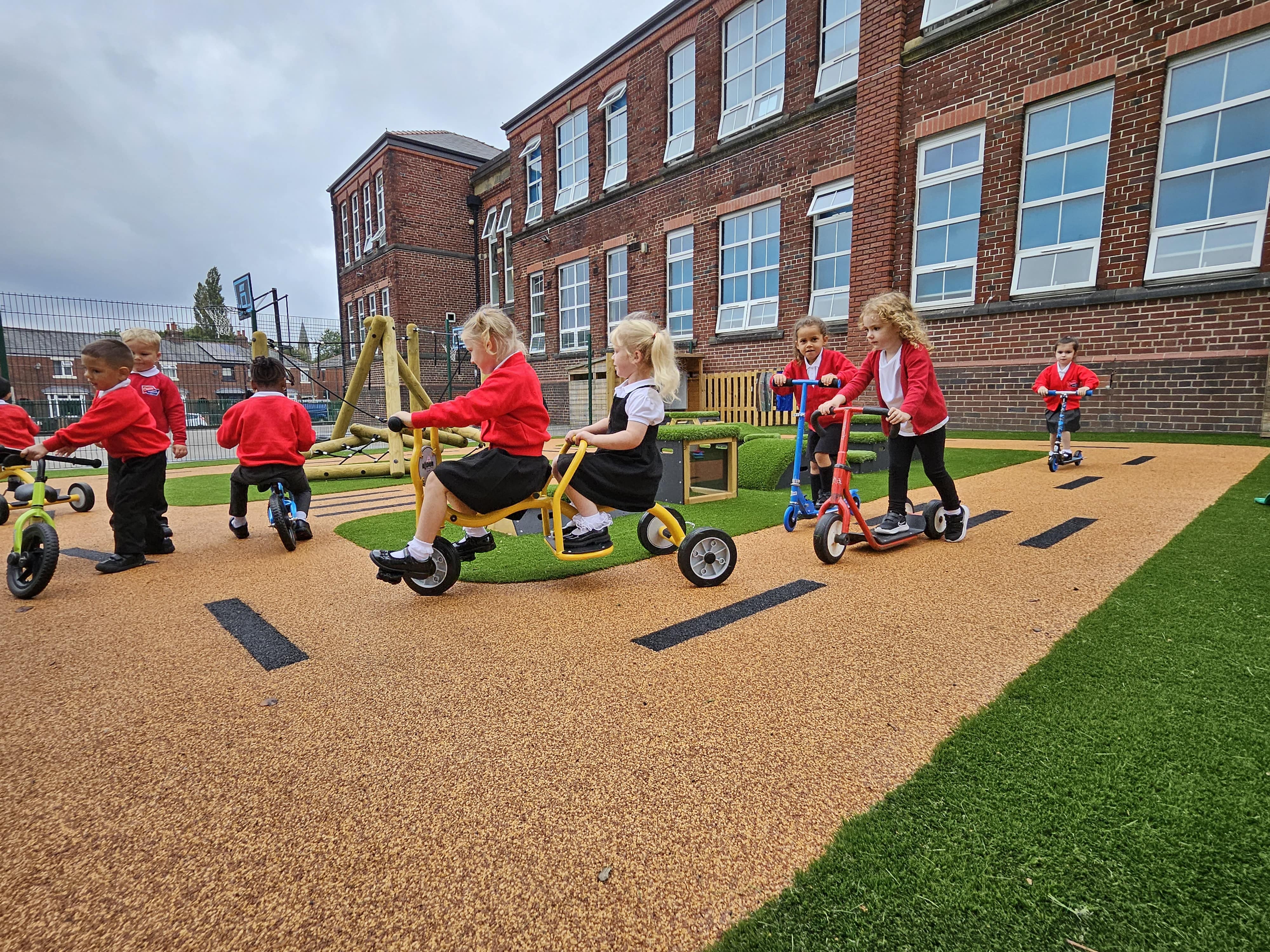 A group of children are playing on bikes and scooters on a wetpour road, with artificial grass surrounding the area. A set of Get Set Go Blocks and a Playframe can be seen on the area.