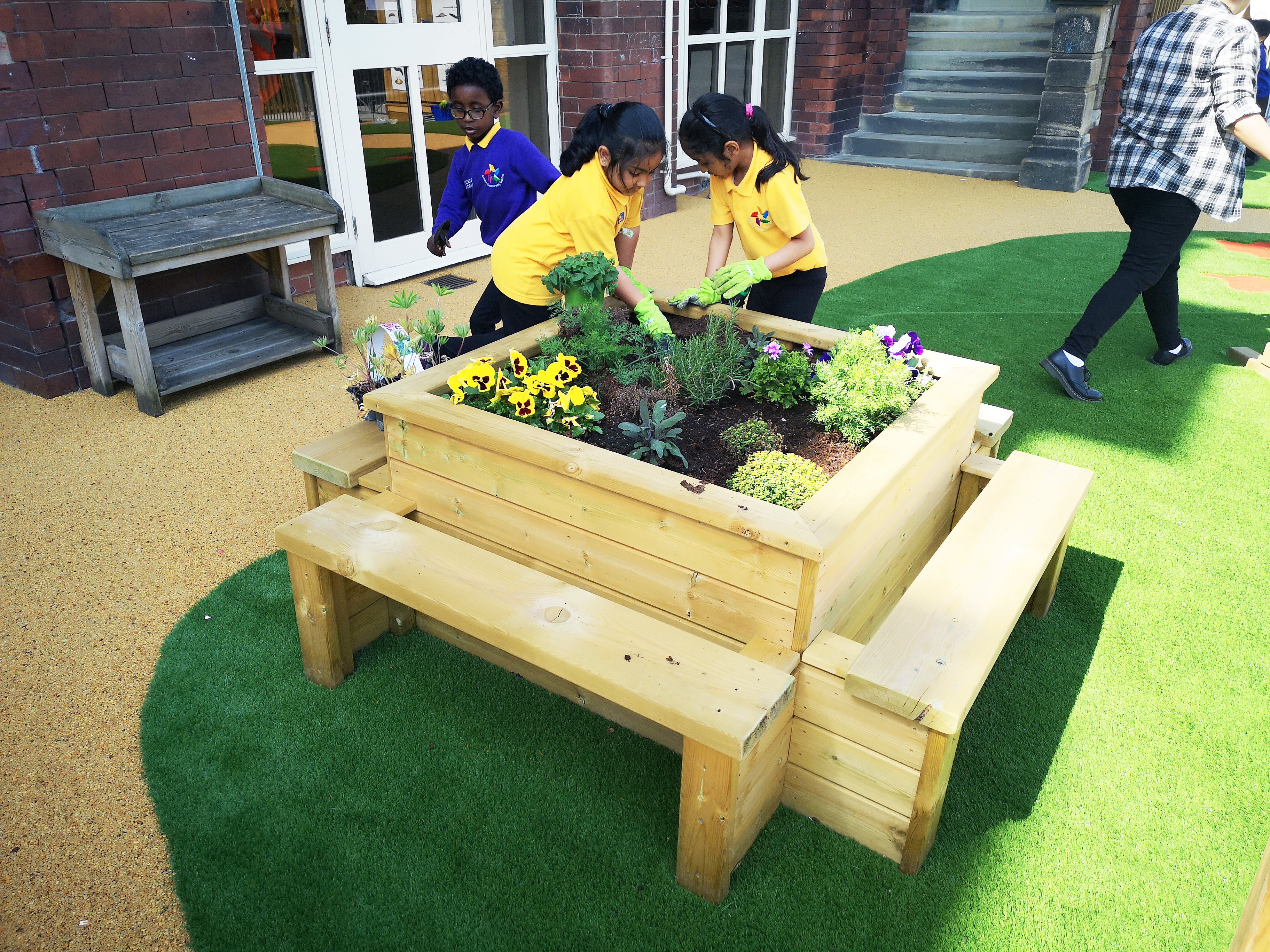 2 children are planting flowers in a square planter. The planter is located just outside the school with it being installed on an artificial grass surface.