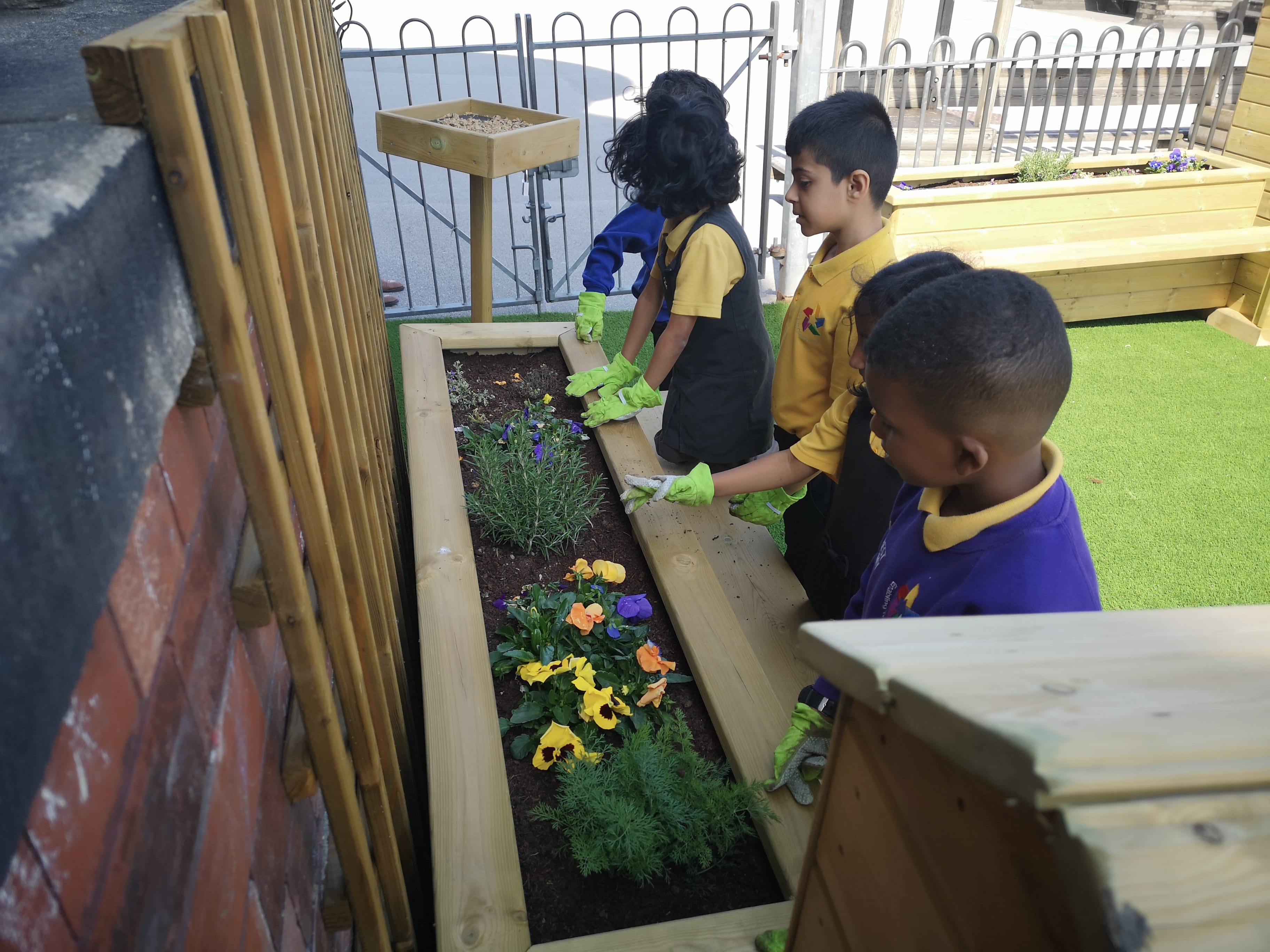 Five children are facing a straight planter and are planting a variety of new flowers into the equipment. The kids are wearing gloves as they talk to each other. The planter is located on top of artificial grass.