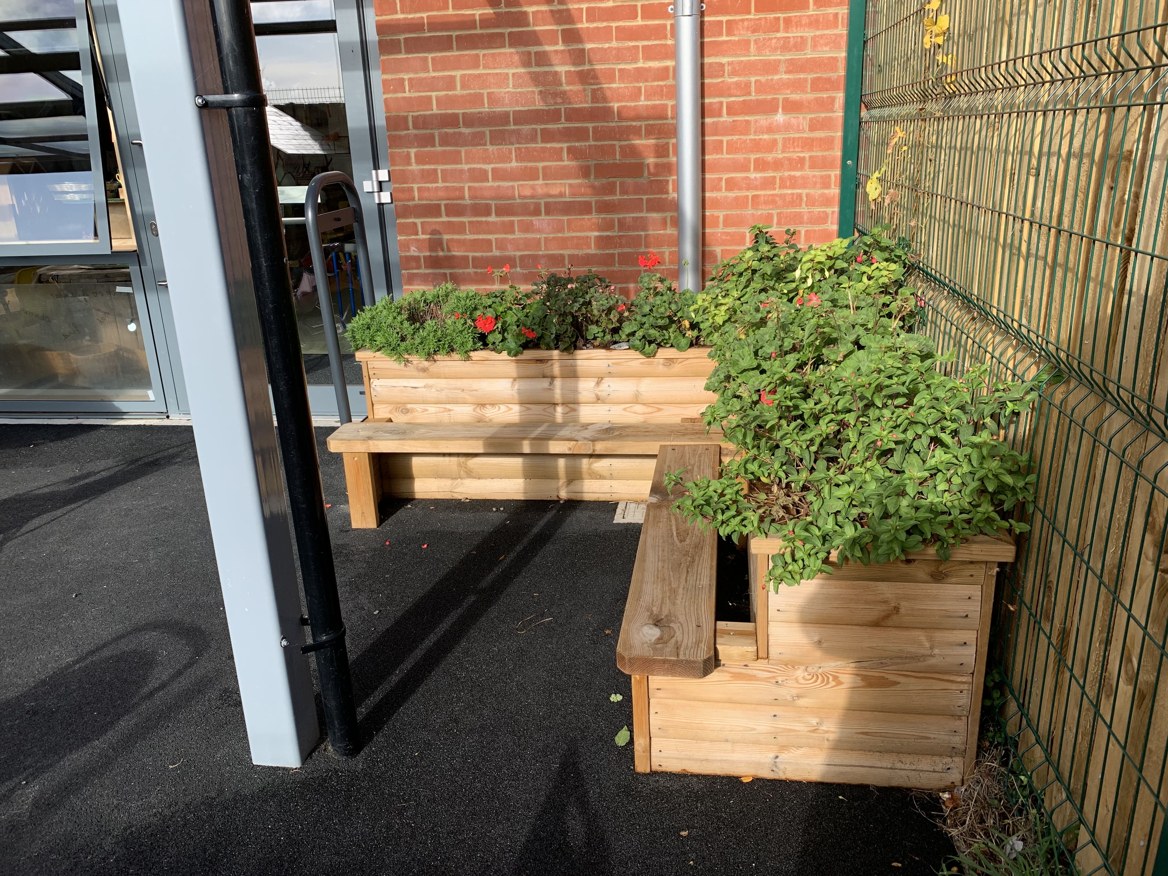 A planter that has been built to be placed into corners with it's right-angle design. The planter has green and red shrubbery planted in it, with benches attached to the actual planter.