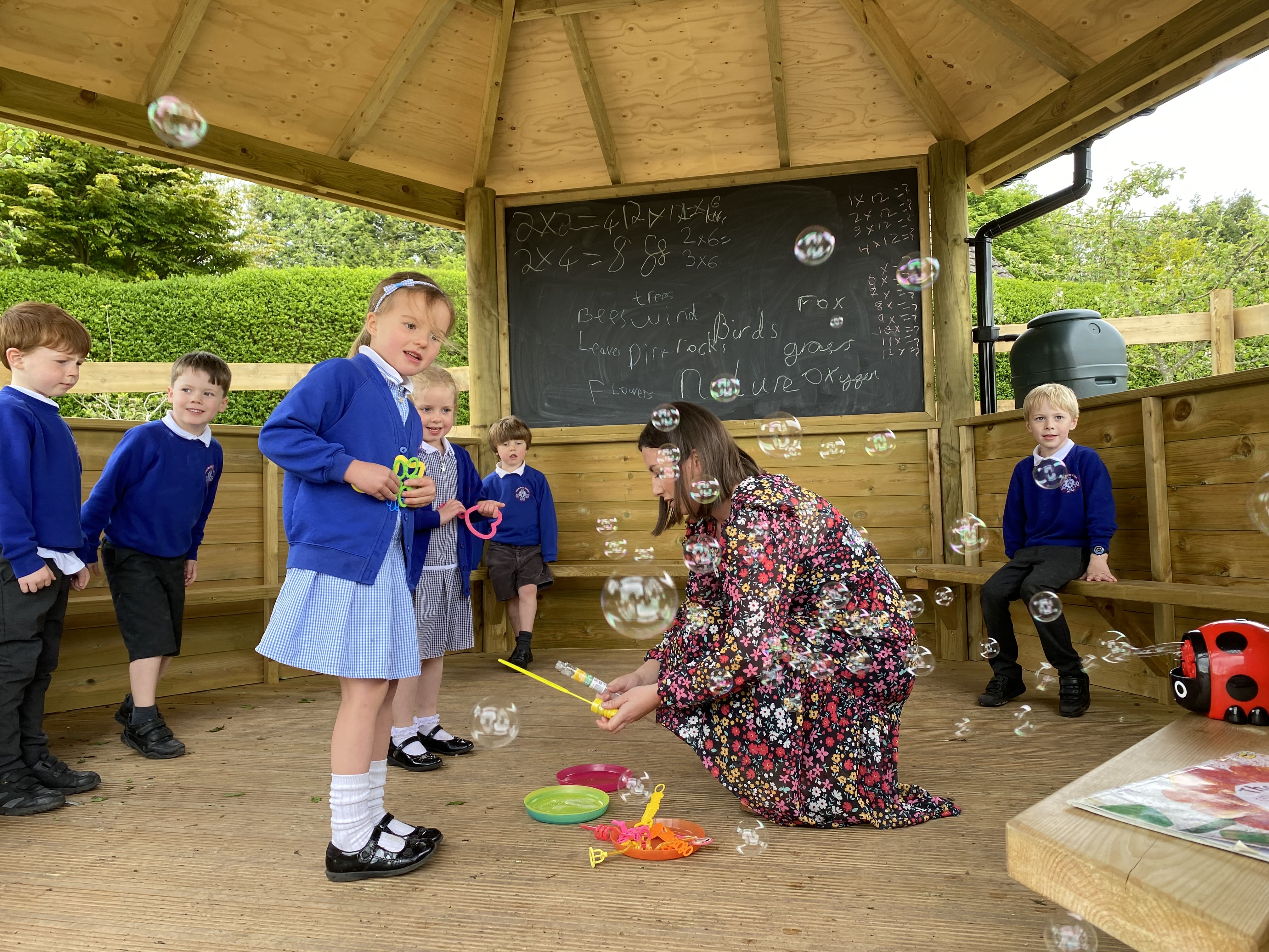 A group of children are playing inside a secret garden gazebo with a teacher and bubbles. A chalkboard has been written on in the background.