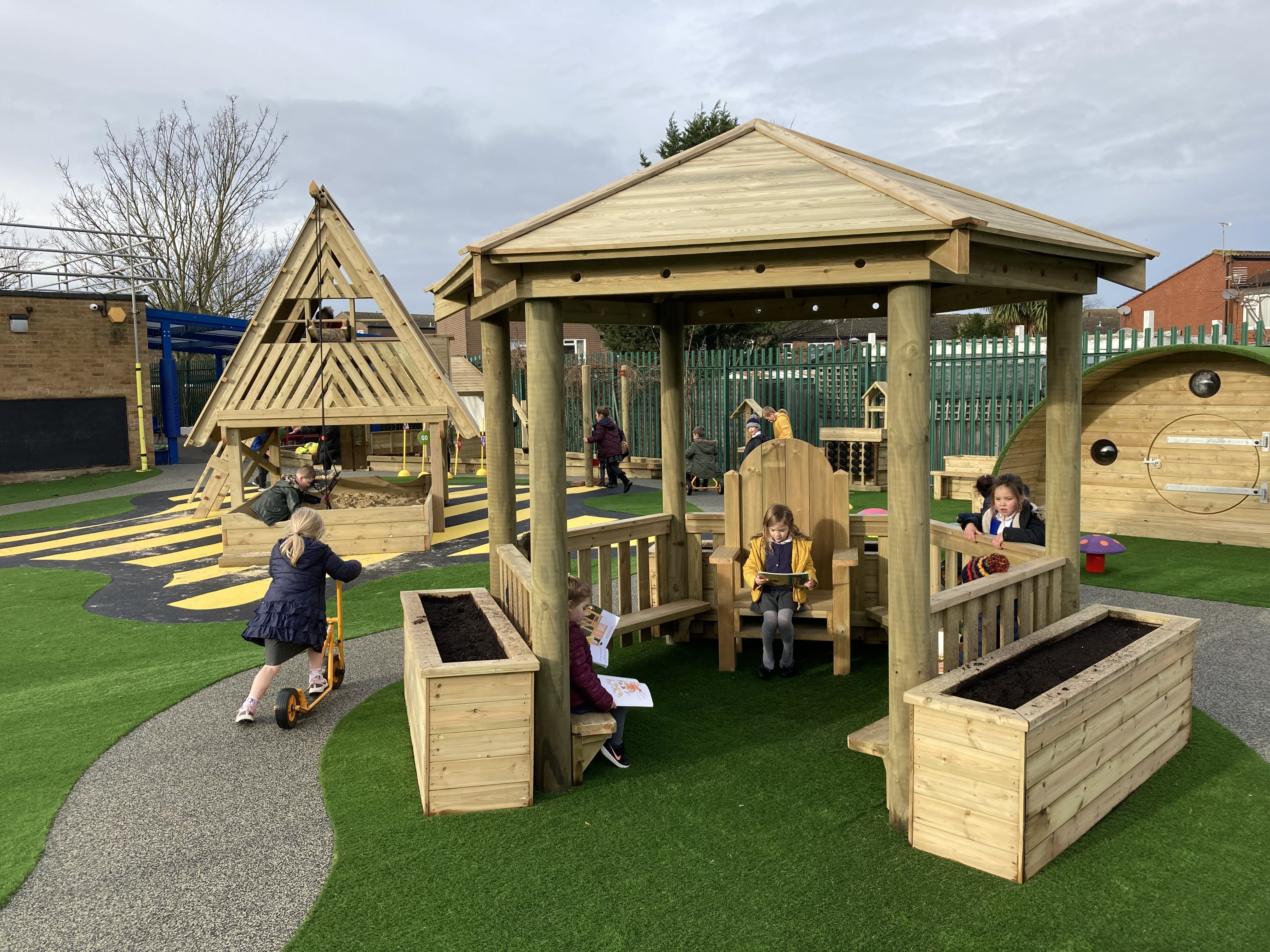 A group of children are underneath the 3.5m gazebo, which has been installed on artificial grass. A wetpour road is going around the gazebo, creating a variety of surfacing to experience. 2 Planters can be seen on the image and located next to the gazebo.