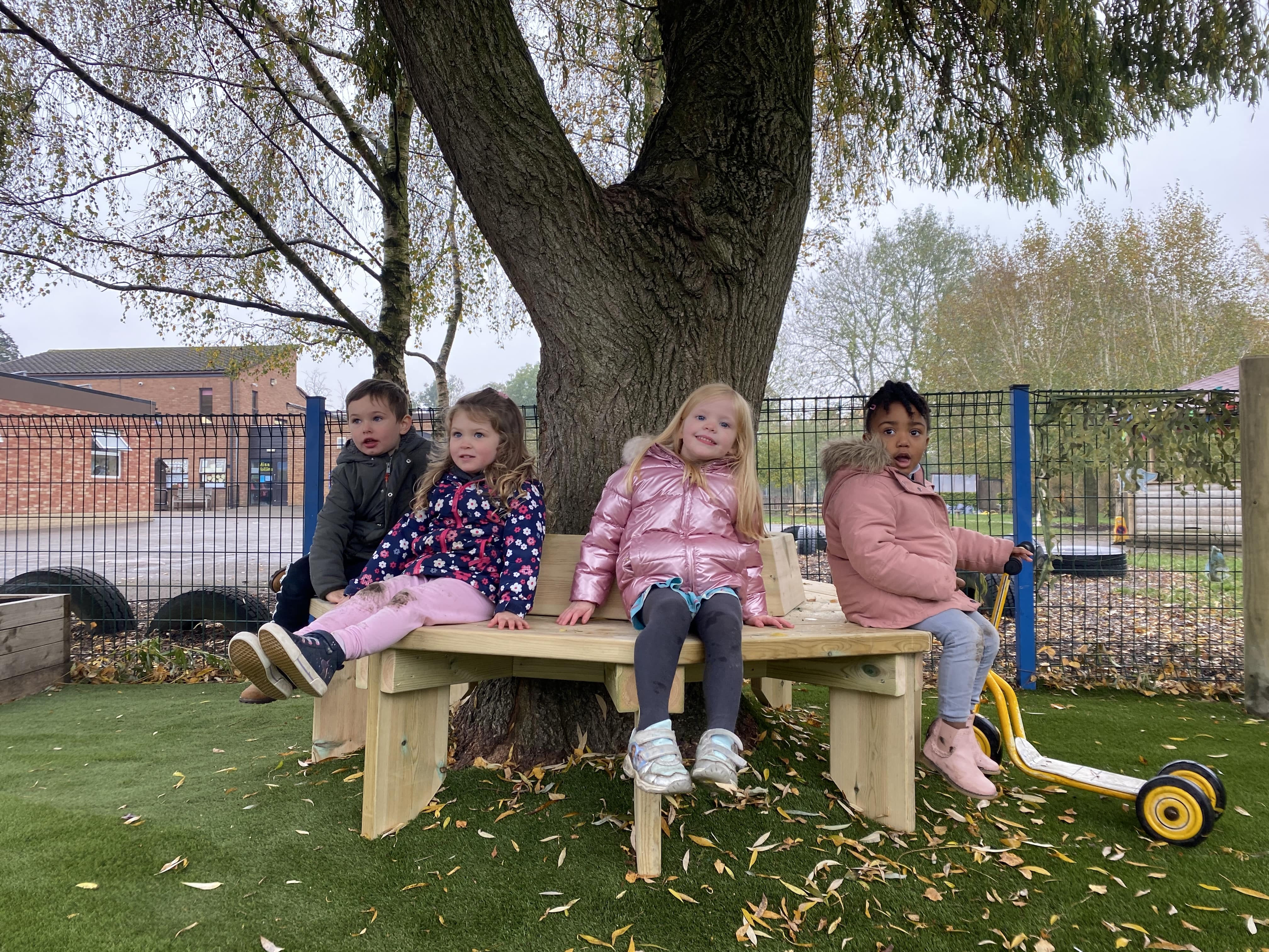 4 children are sat on a wooden bench that goes around a big tree. The children are sat at the bench and are looking happy as they look towards the camera.