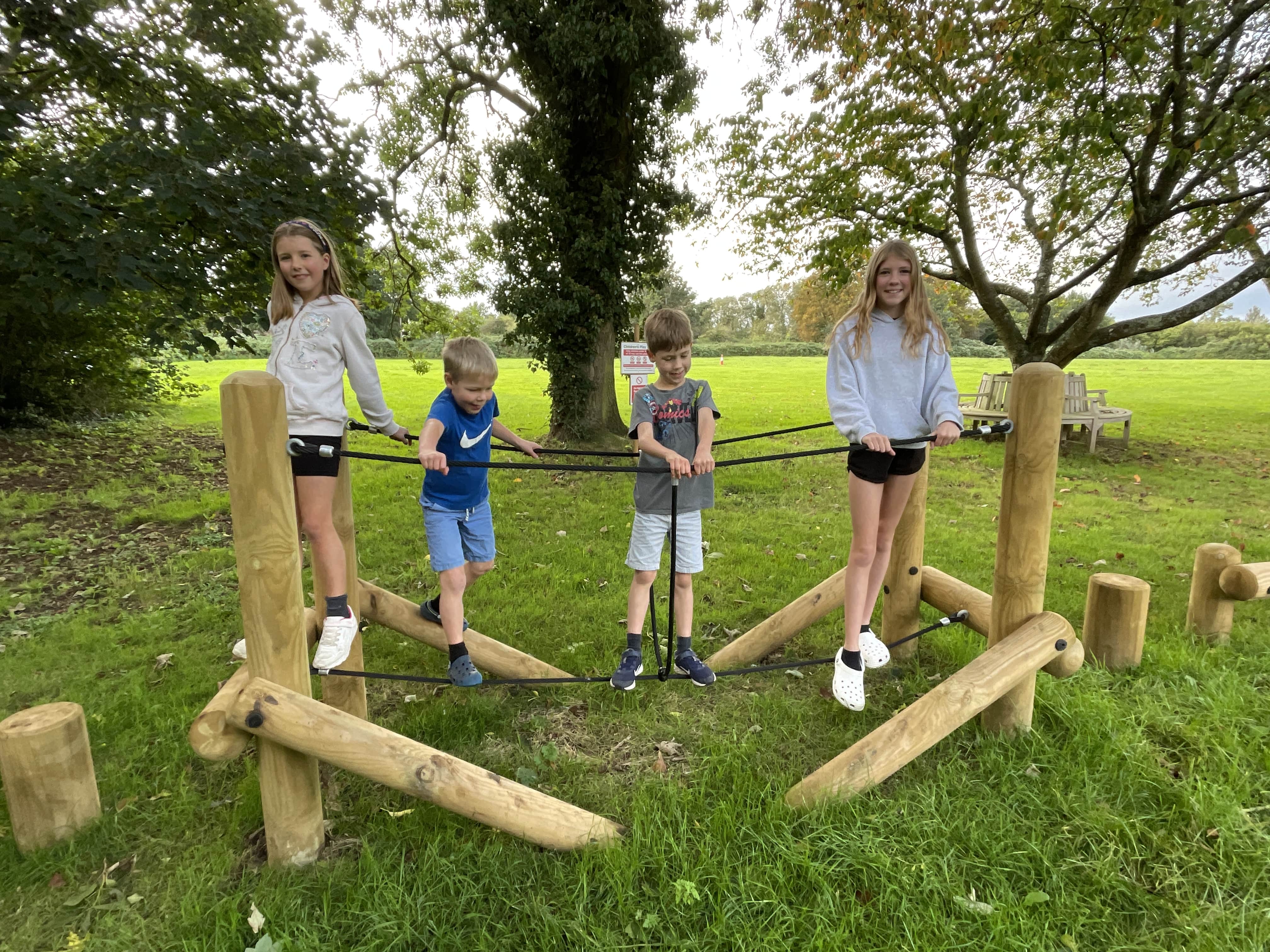 4 children are balancing on the tightrope bridge as they look at the camera and smile. The tightrope bridge has been installed on natural turf and surrounding trees.