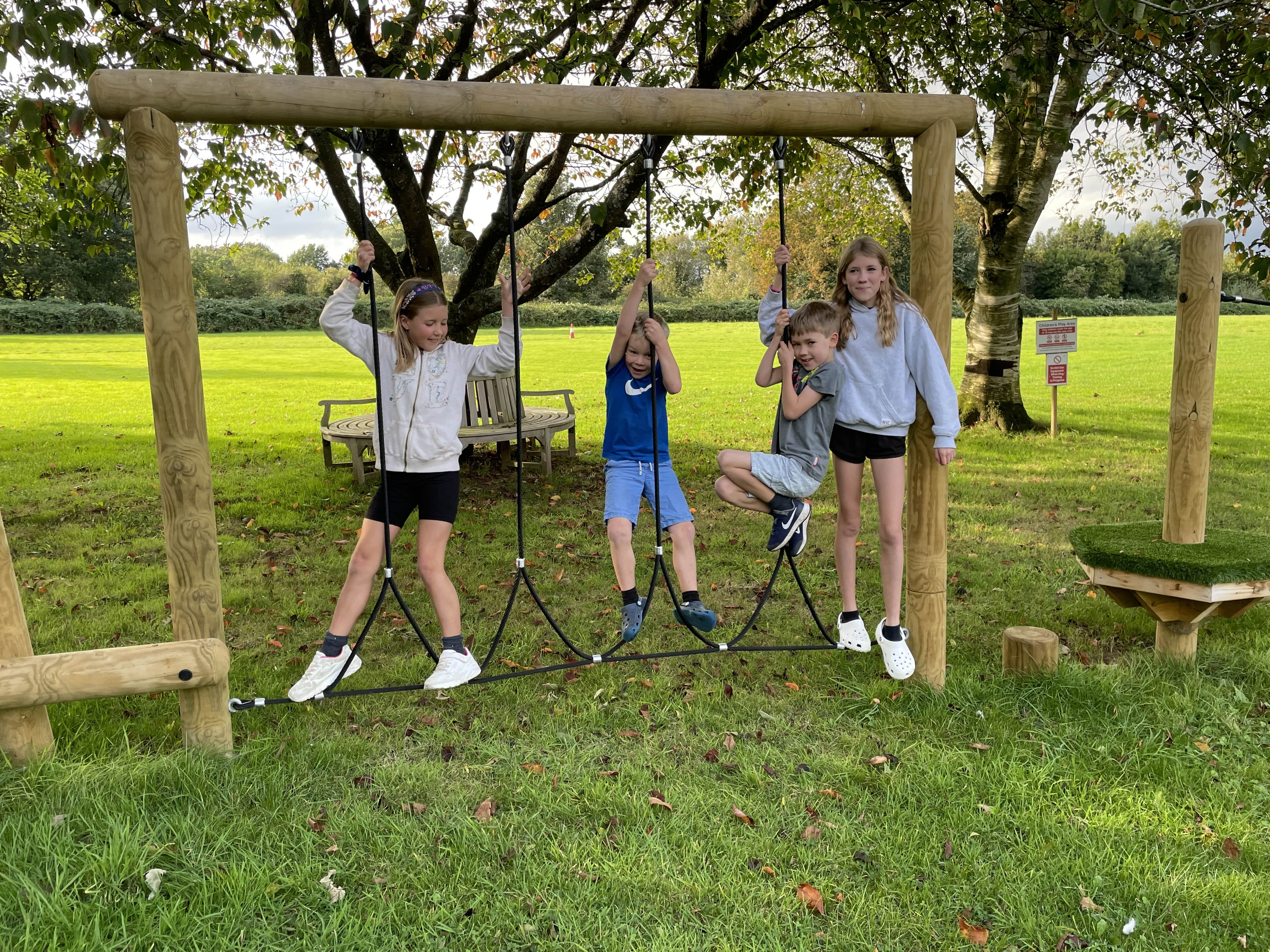 A group of 4 children are playing on the step rope traverse piece of equipment. The kids are smiling and looking in all directions as 2 trees can be seen behind them with two pieces of play equipment on either side.