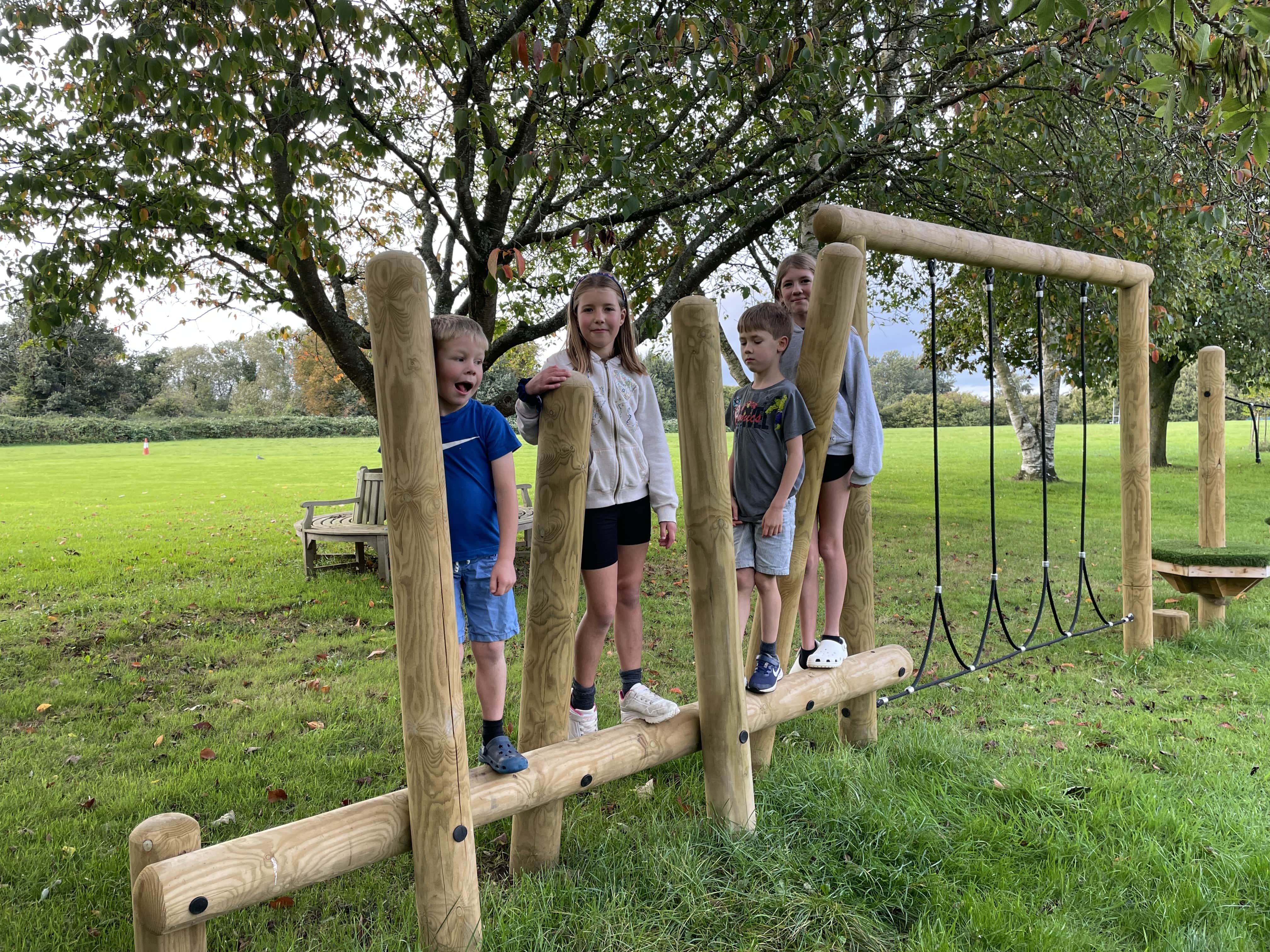 4 children are stood on the balancing beam and are looking at the camera and smiling. You can see the rope traversal equipment, with a big tree and bench behind them.