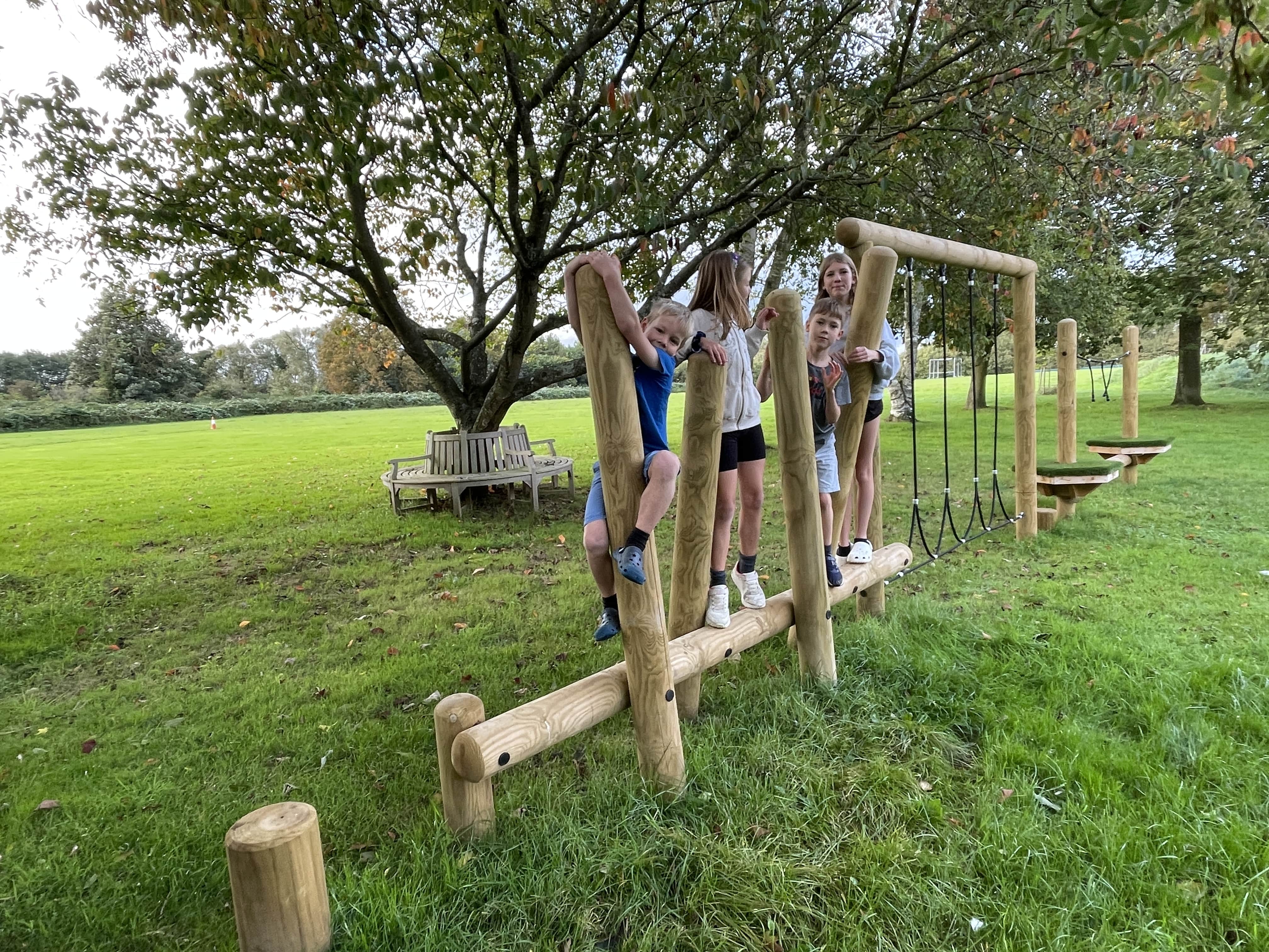 Kids are crossing the wooden balance beam as they hold on to other wooden poles for support. All 4 of the children are smiling and having fun as they navigate the obstacle course. Trees and natural grass can be seen surrounding the area..