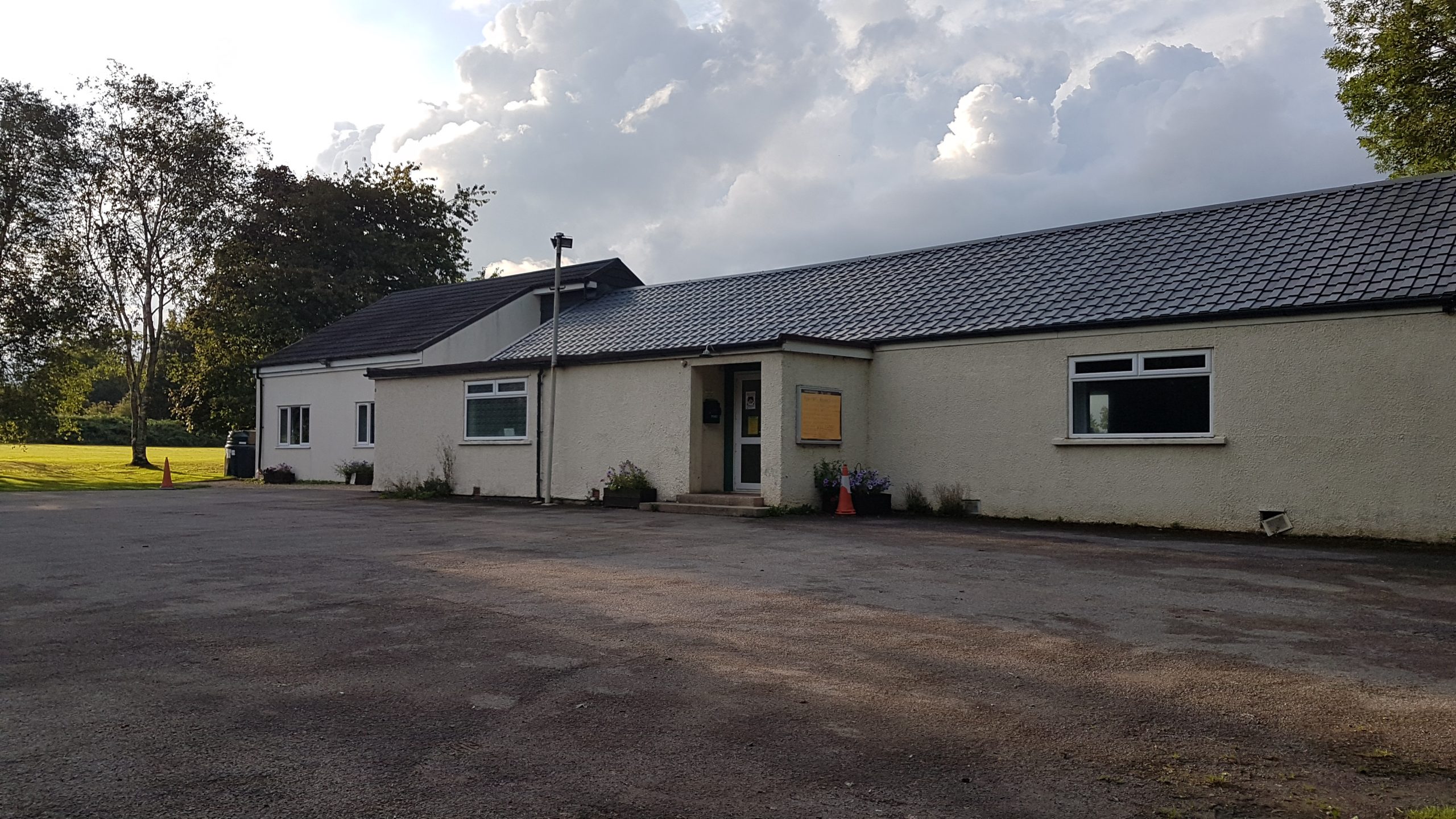 The outdoors of the Village Hall in Langley Burrell. It's a white bungalow shaped building with a concrete car park with a grass area to the side.