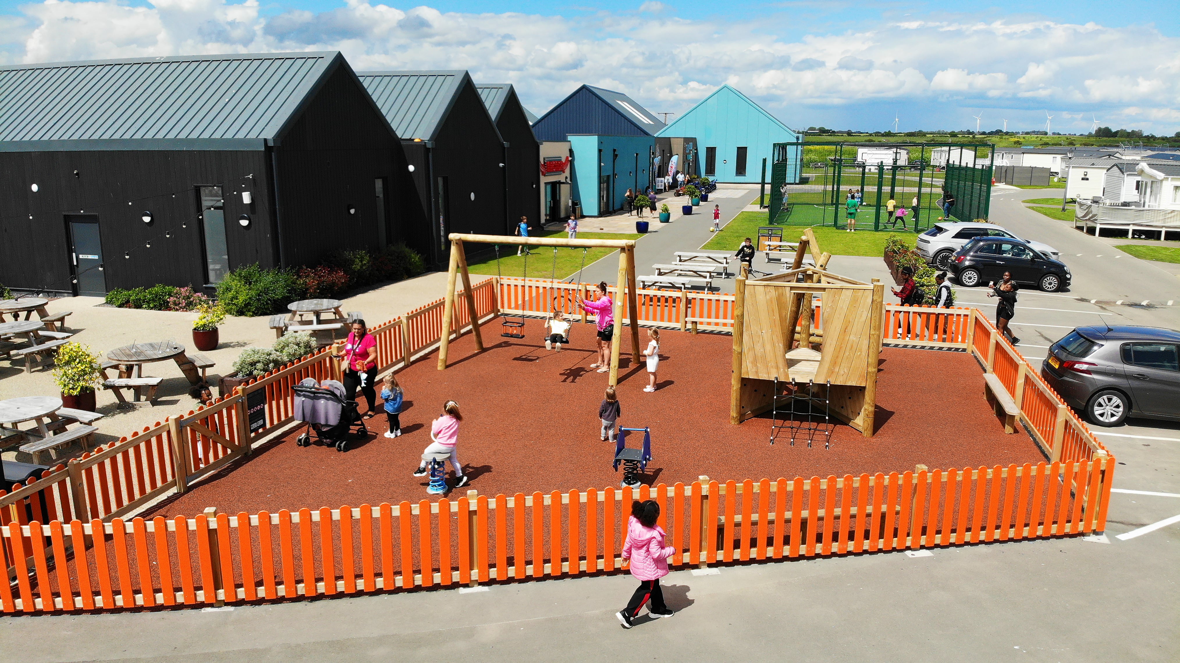 The orange wetpour playground is surrounded by orange HDPE Bow Top Fencing. A large group of people can be seen playing on the park.