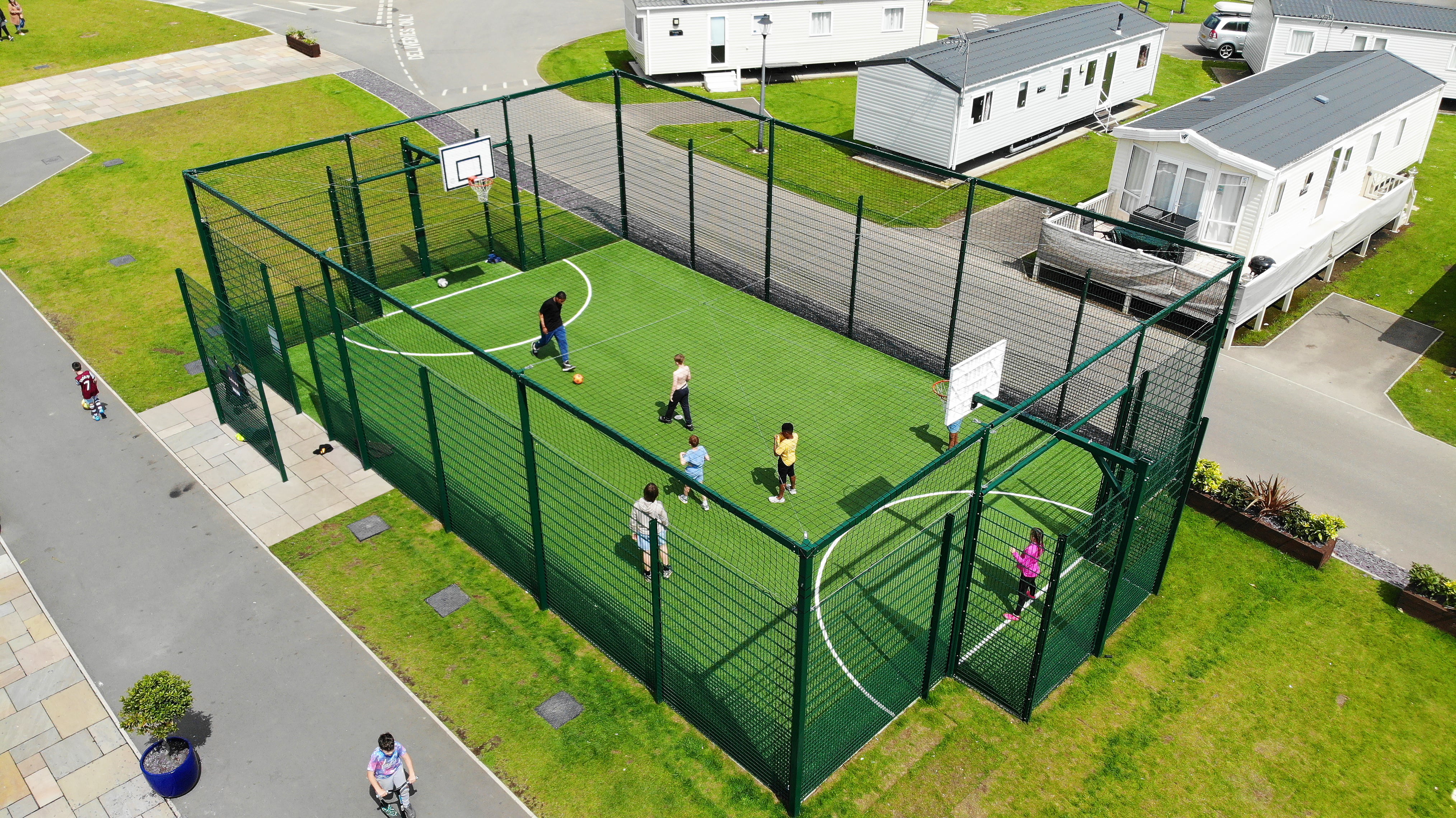 A MUGA with a 4g artificial grass surfacing, with line markings. The MUGA is surrounded by green metal fencing and has basketball posts and football goals built into it's design.