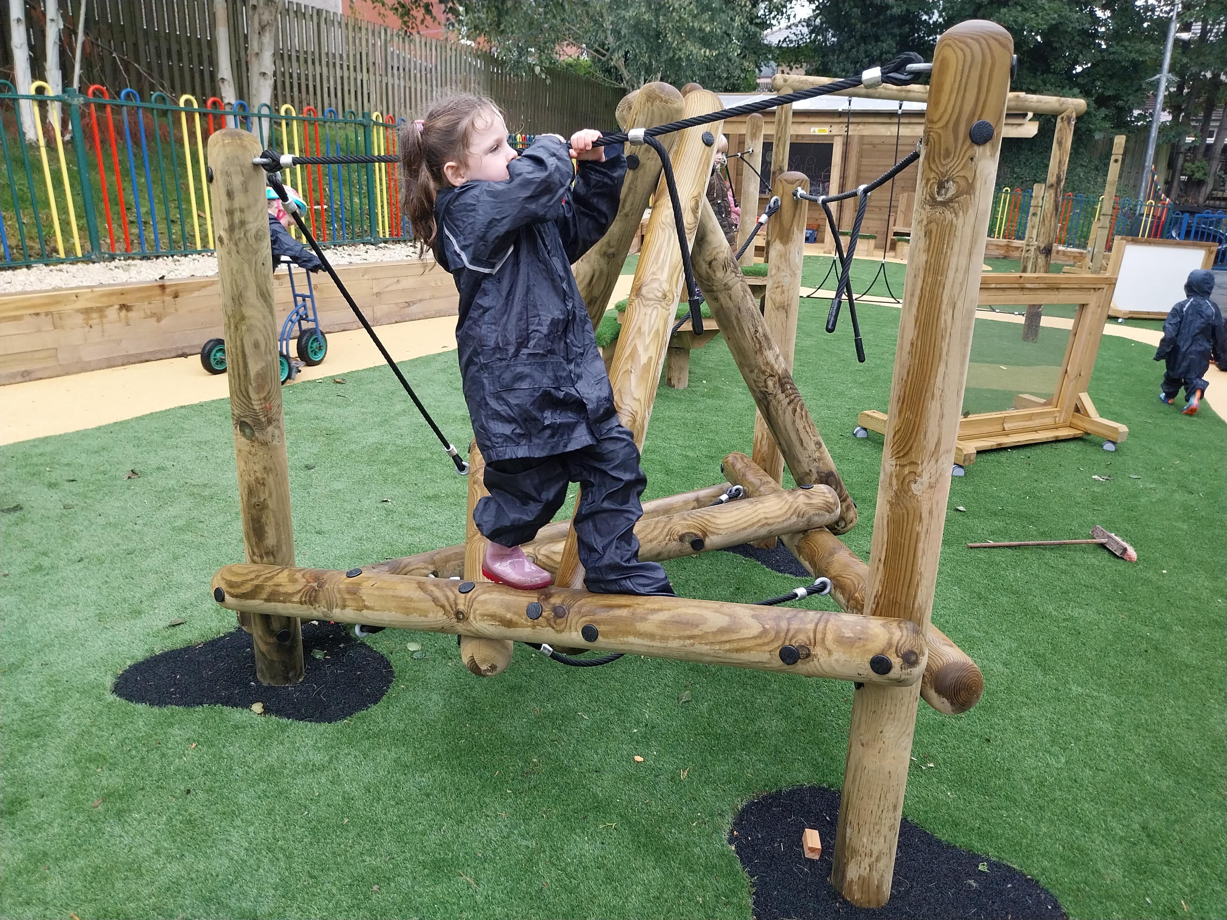 A child is playing on the Pinnacle Hill Climber as they hold on to the rope. They are balancing their way across the beam. Artificial grass surfacing surrounds the play equipment.