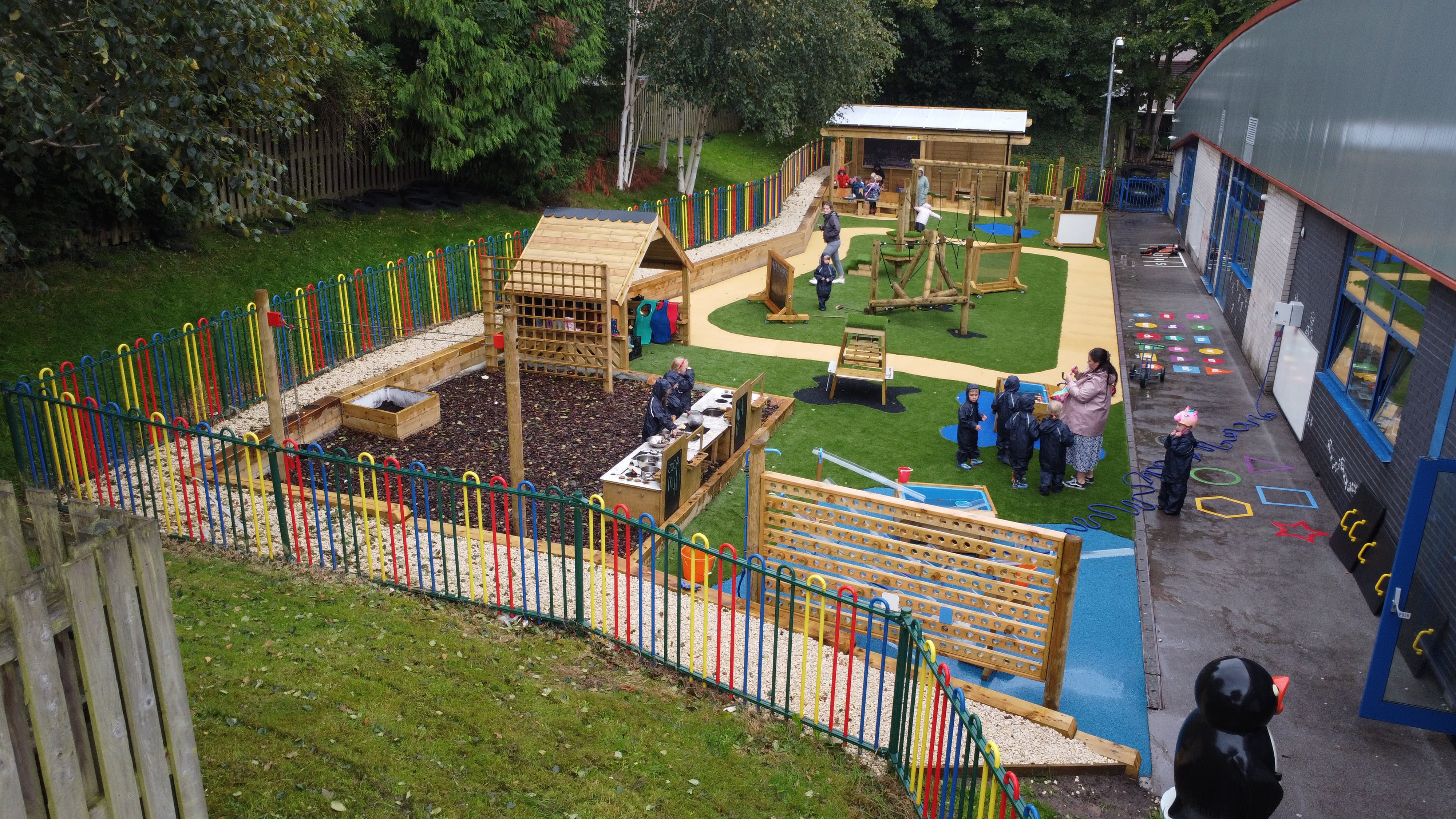 A far away photo showing all of the play equipment on the playground, including the surfacing options that were selected. A large group of children can be seen playing on it and enjoying themselves.