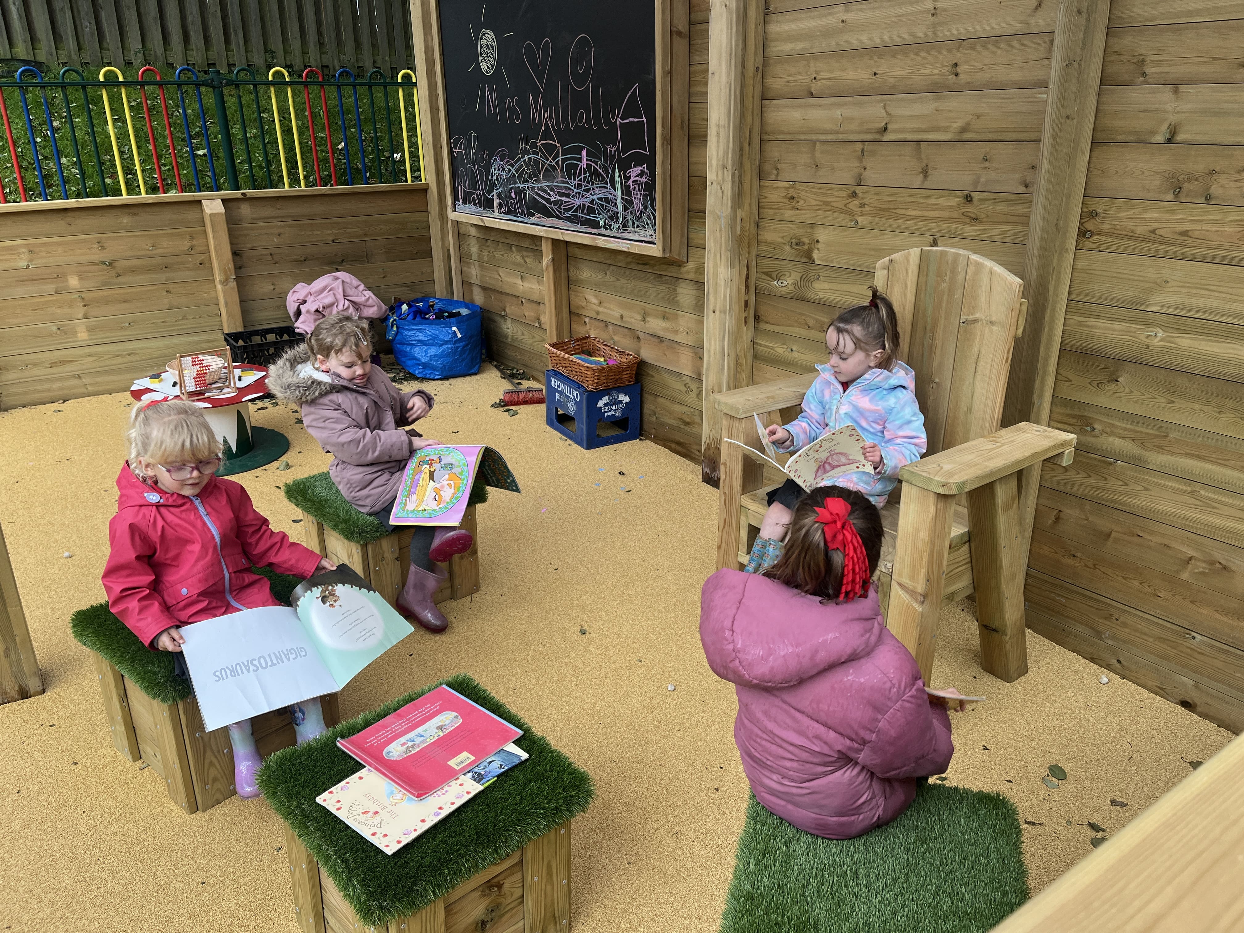 A little girl is sat in a freestanding storytelling chair as she reads a book. Three other children are sat on artificial grass topped seats opposite her and are reading books.