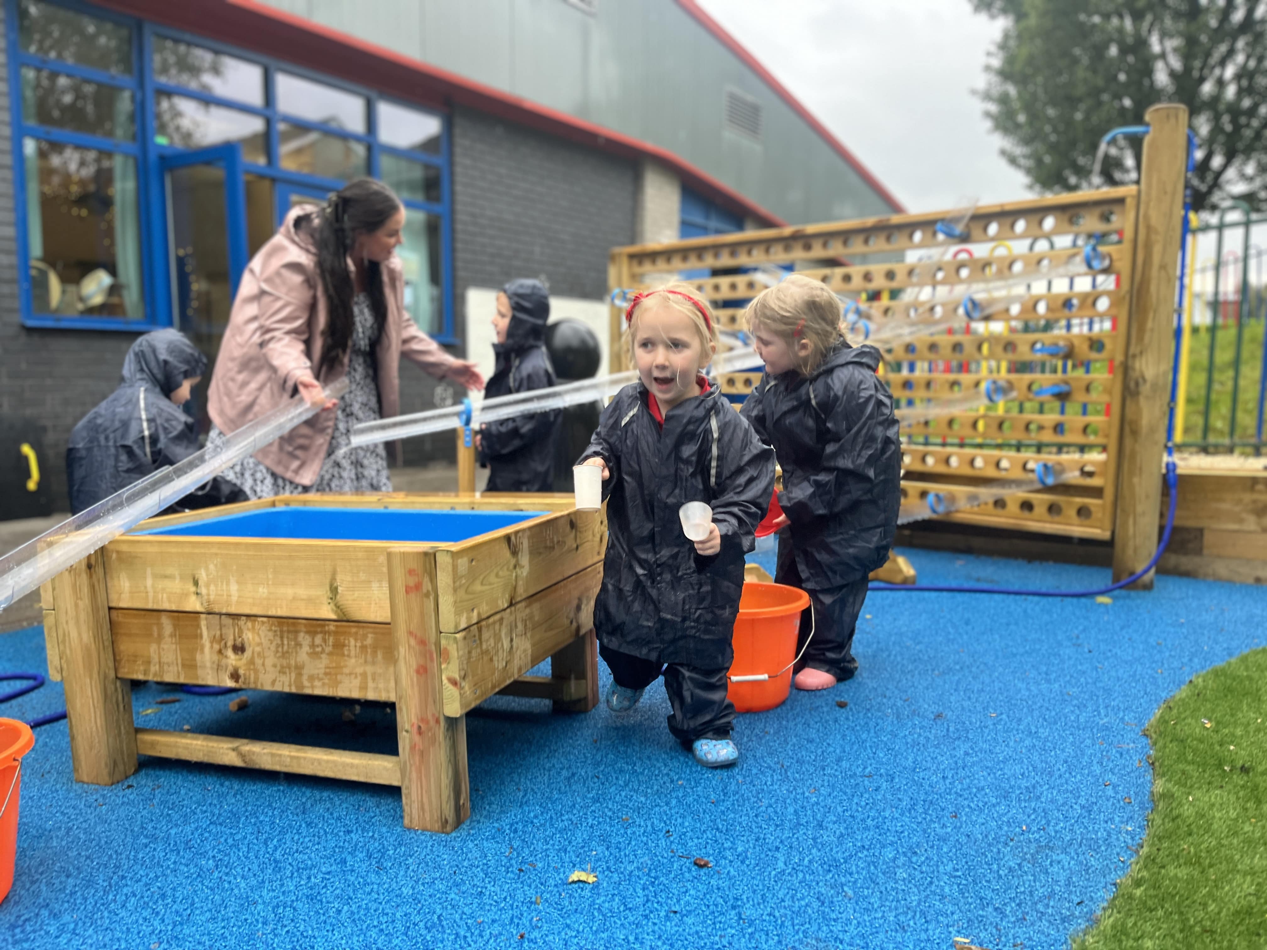 A little girl is running towards the camera and carrying some water play equipment, as she smiles and cheers. The teacher and other children are having a fun time learning about water.