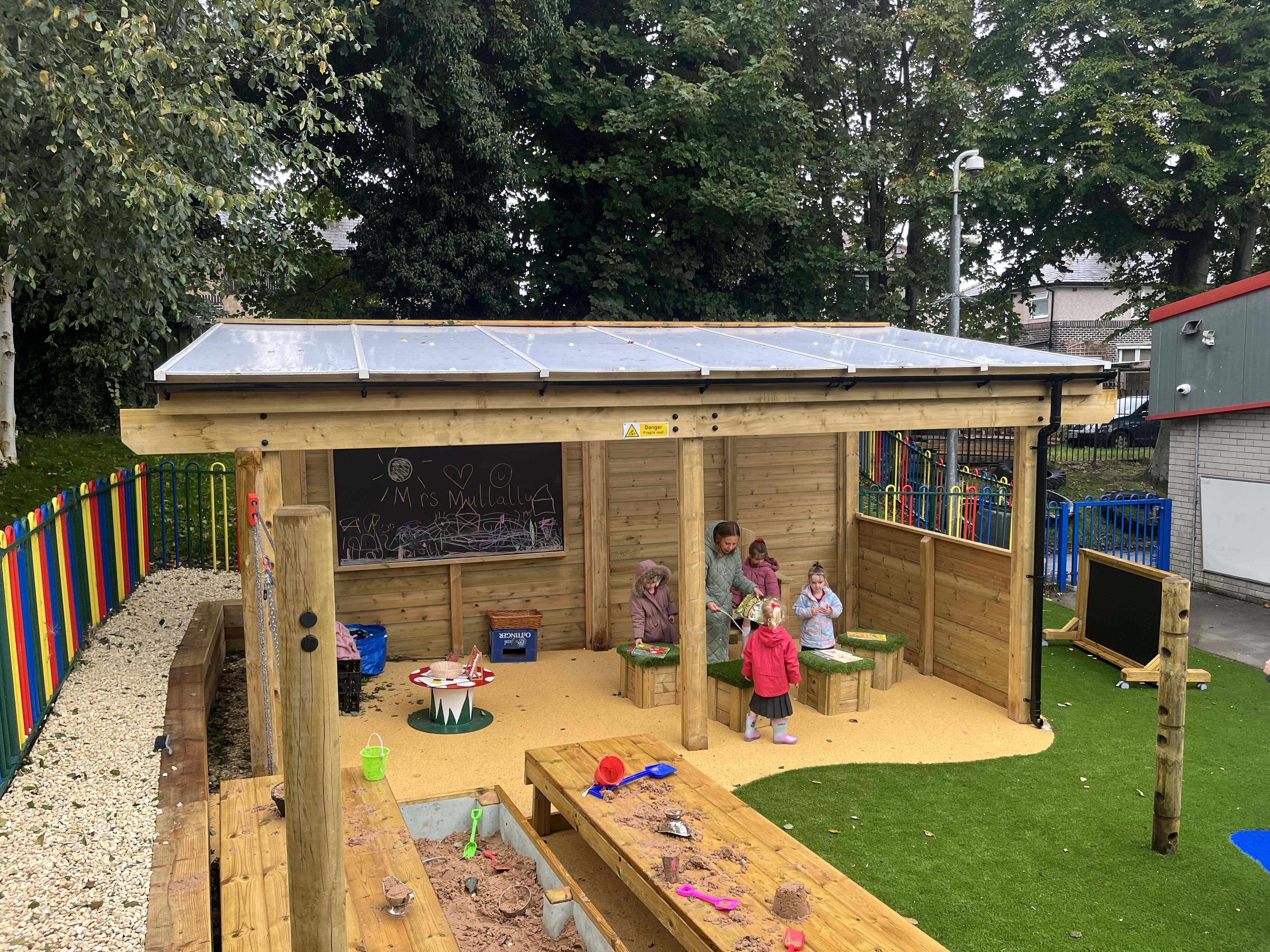 A timber, freestanding canopy that has been installed on a playground. The children are sat in the canopy as they take part in an outdoor lesson.