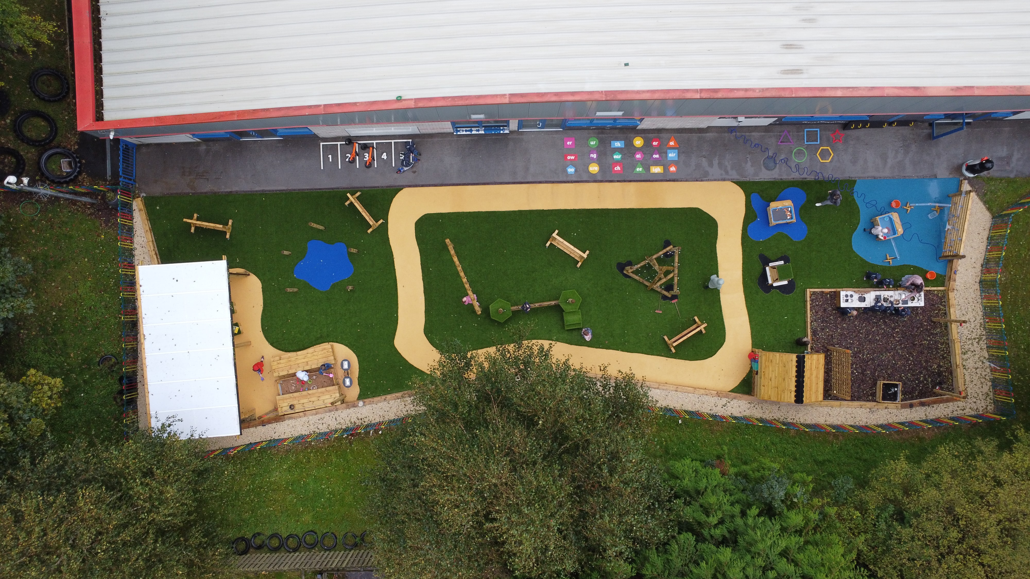 A view of the entire play area surface, showing the artificial grass surfacing with patches of wetpour installed on it. Next to the artificial grass play area, a tarmac area can be seen with thermoplastic markings.