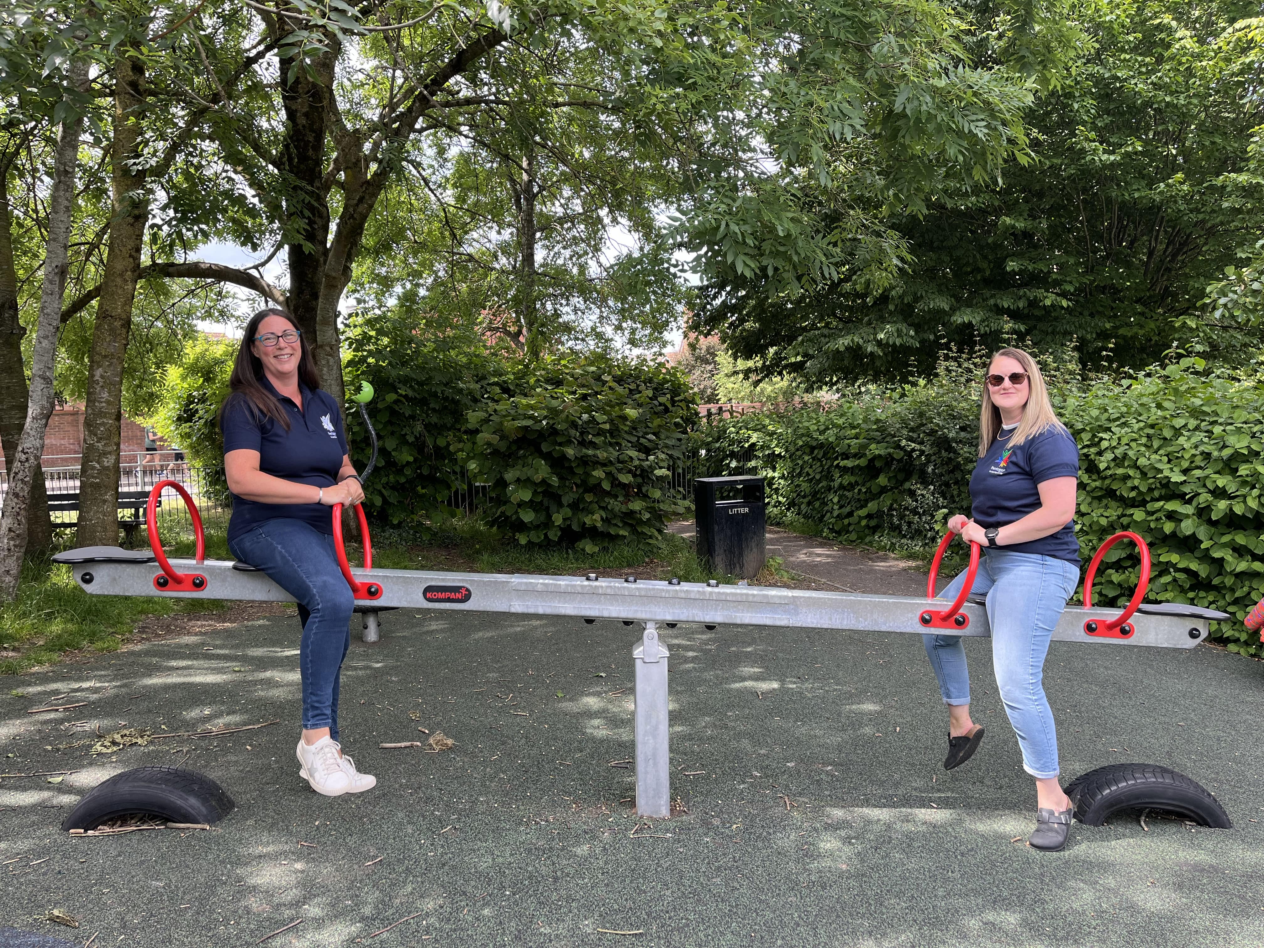 Two Pentagon Play inspectors sitting on a see saw and are smiling and looking at the camera. The see-saw is placed on top of a wetpour play area.