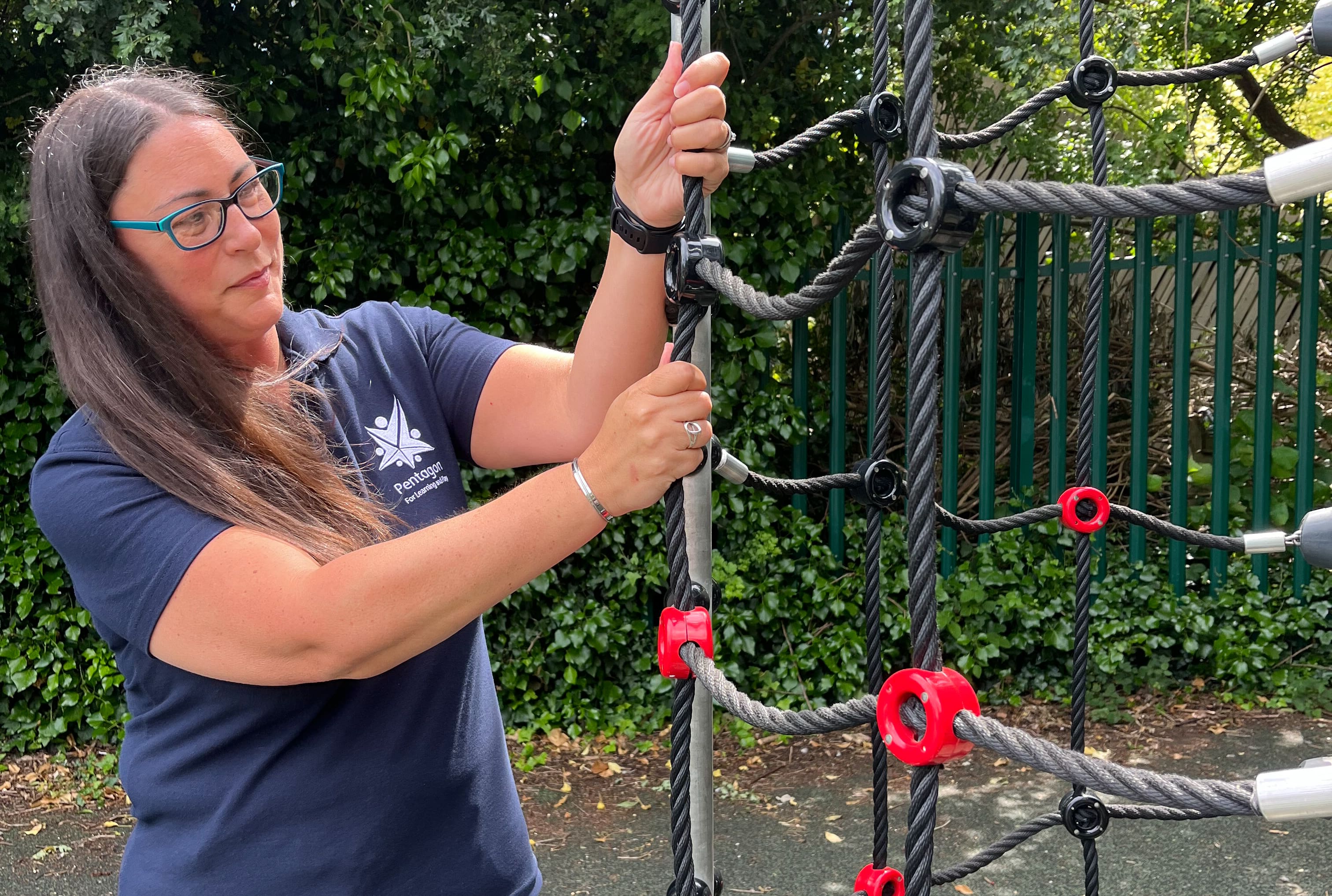 A female playground inspector from Pentagon Play who is inspecting a climbing net. She is grabbing the net and giving it a pull.