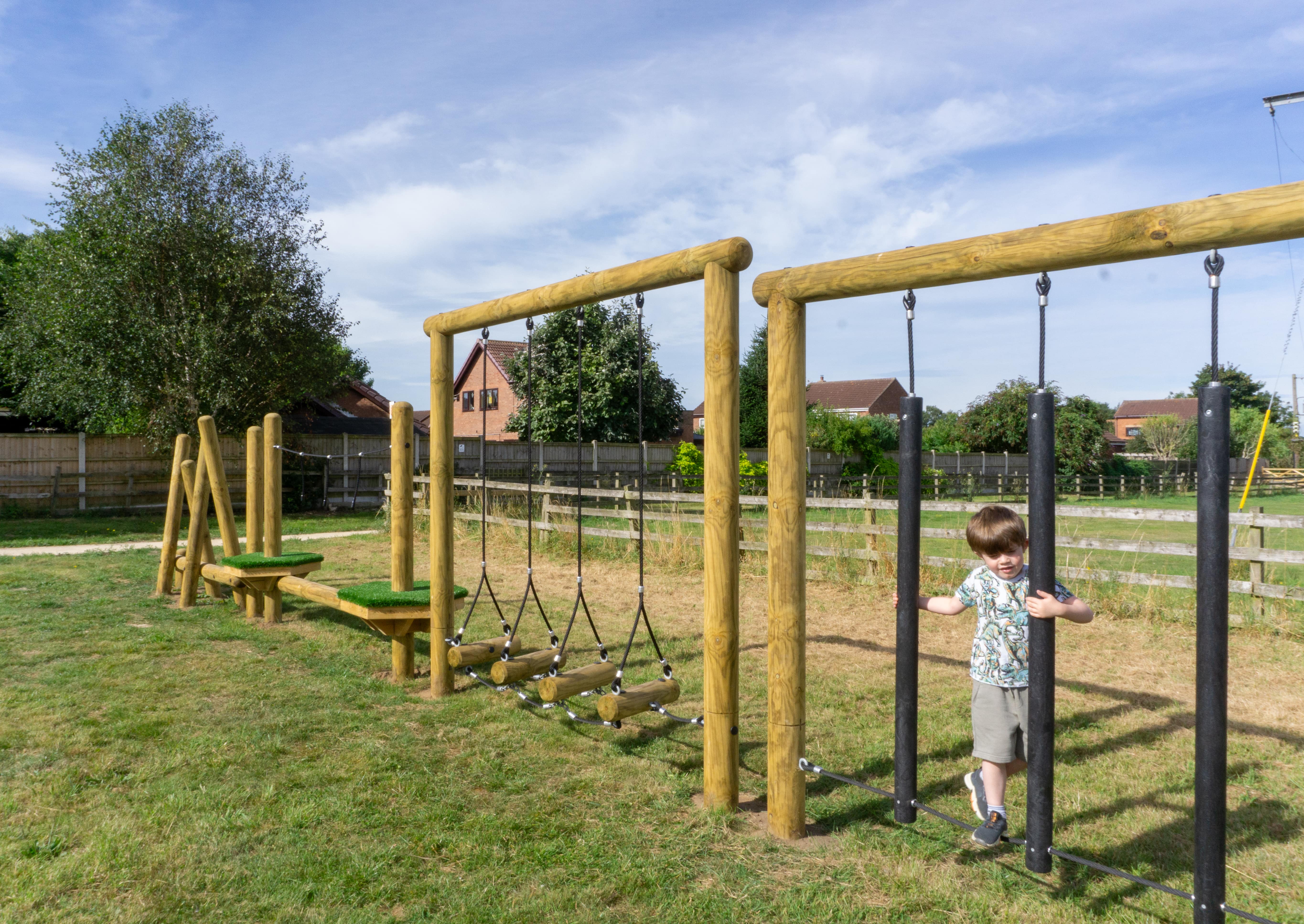 A child is on a rope traversal piece of equipment and is making his way through the Trim Trail. A variety of different pieces of the Trim Trail can be seen.