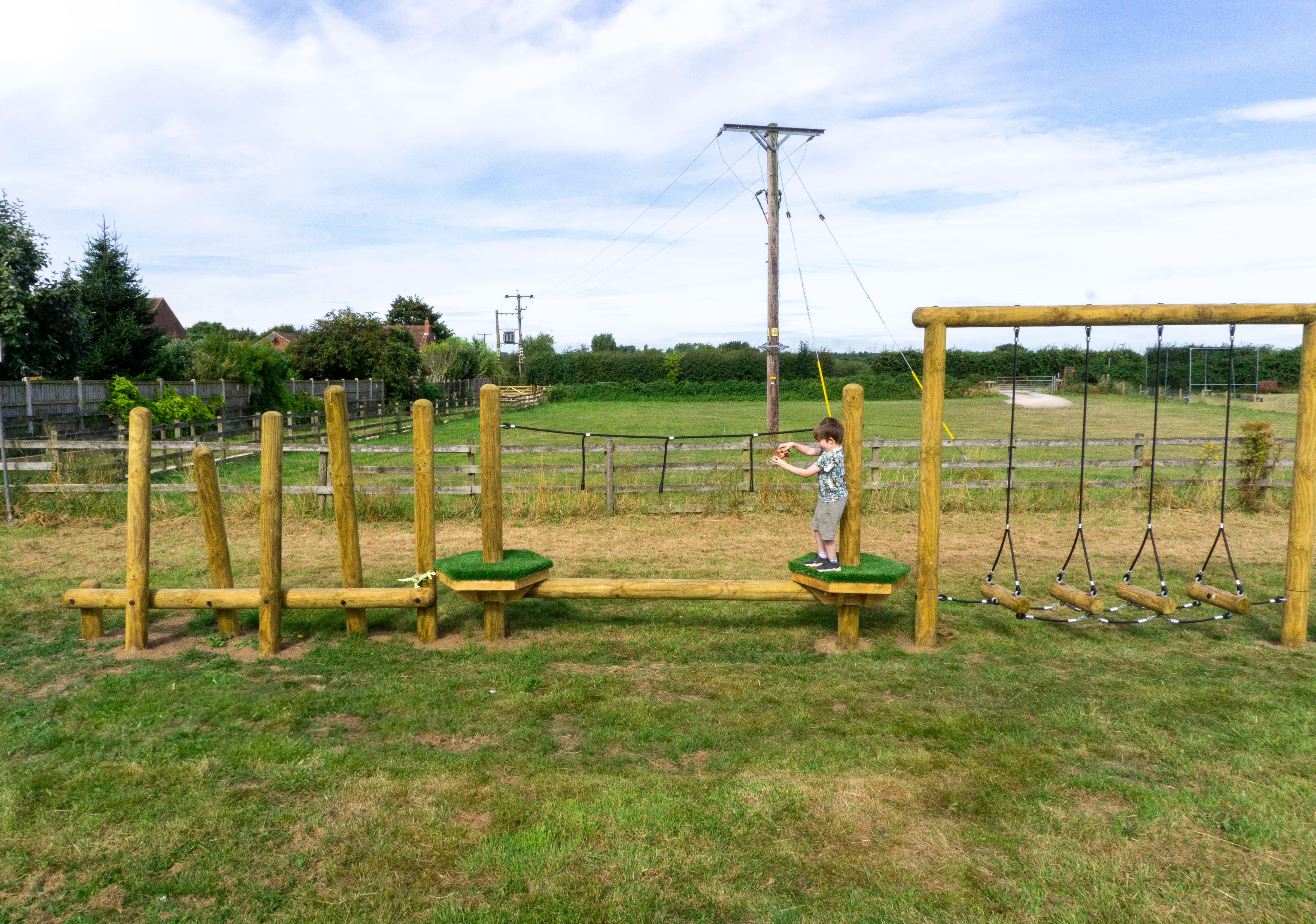 A child is stood on an artificial grass podium and is looking at how he can balance across the wooden beam to make it to the other side.