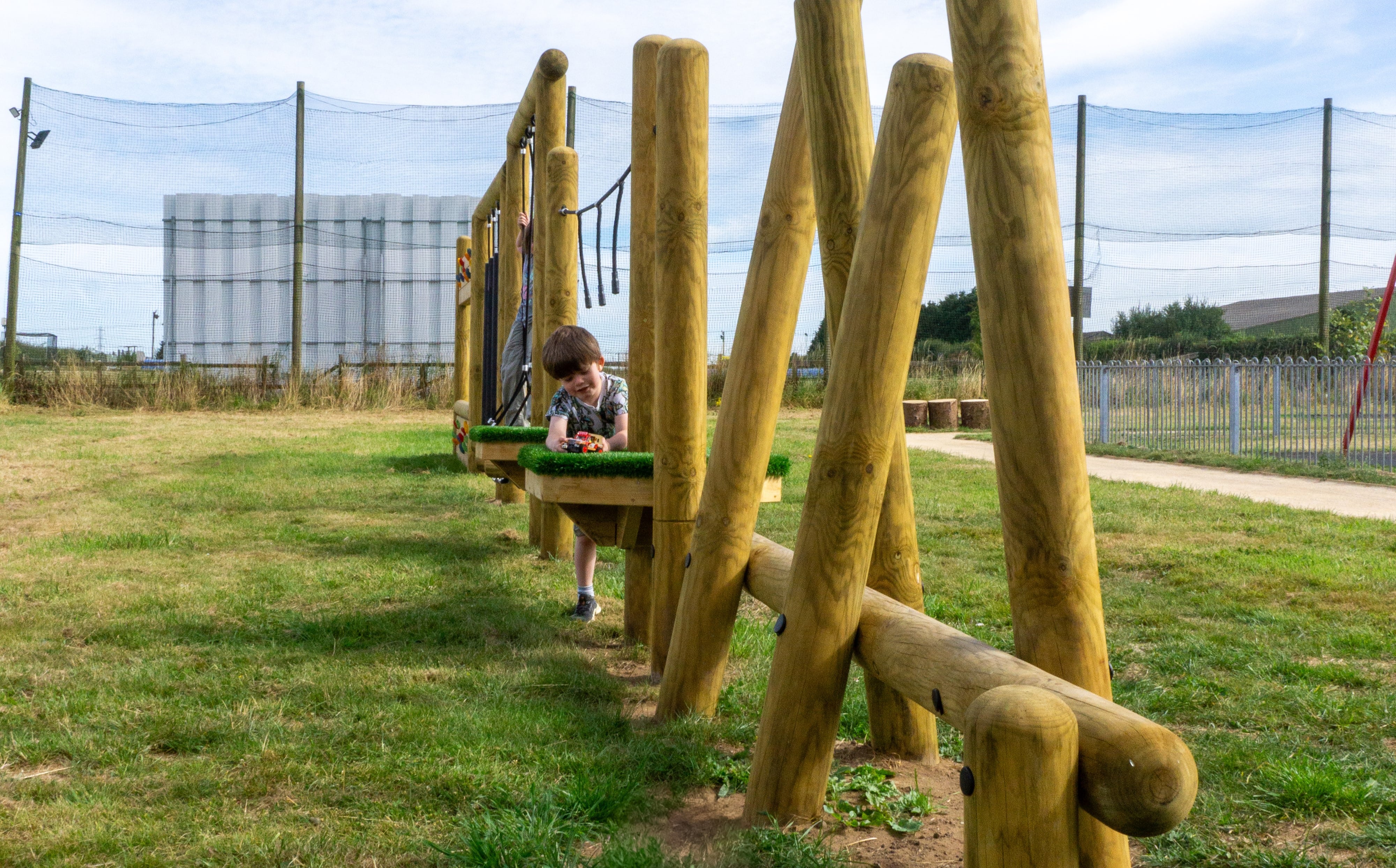 A little boy is stood beside one of the wooden poles and is leaning on an artificial grass podium. He is looking into the camera and is laughing.