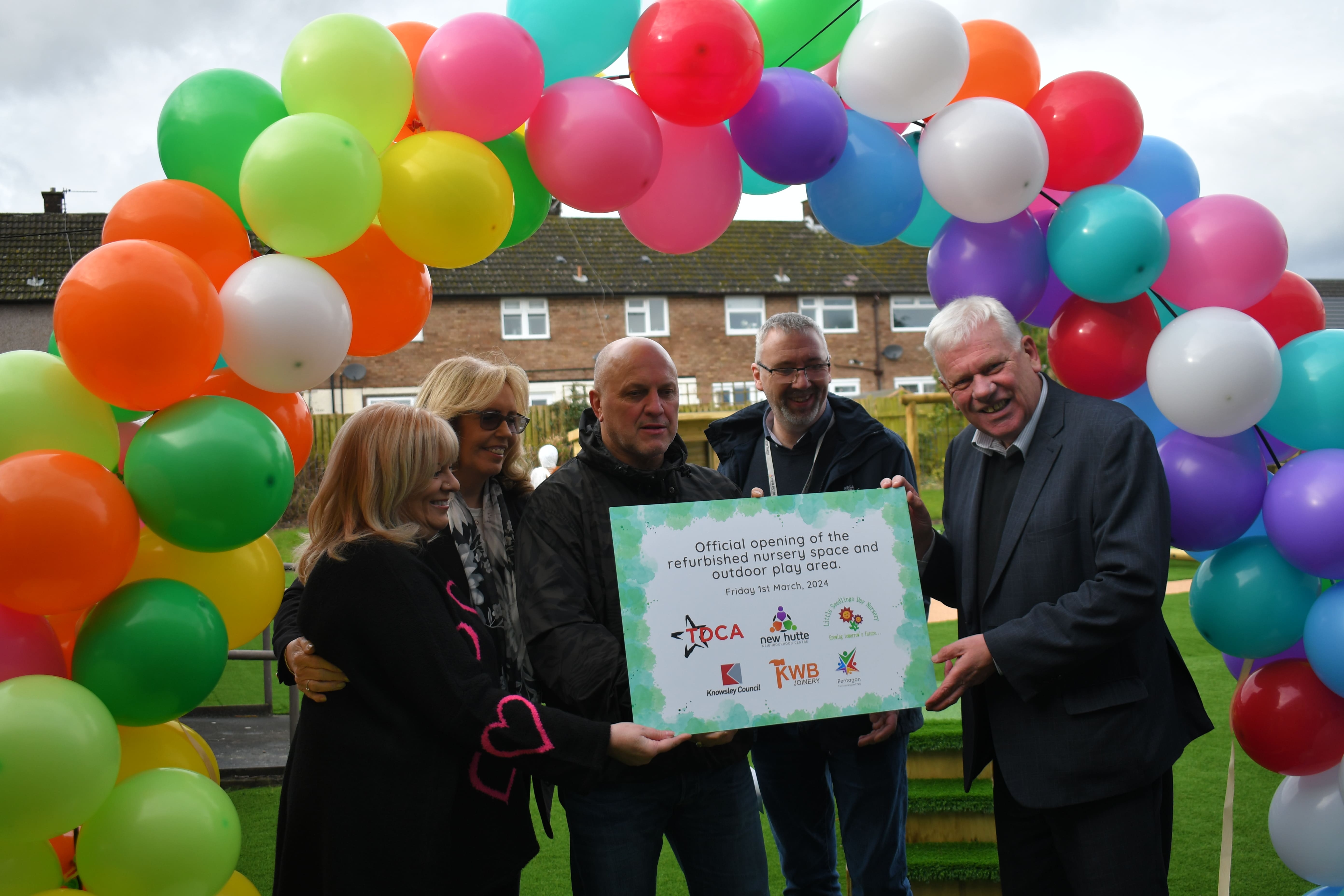 A group of people are stood under a multicoloured balloon arch as they hold up a sign. The sign says "Official opening of the refurbished nursery space and outdoor play area. Friday 1st March 2024." It also has a variety of logos on it, one being Pentagon Play.