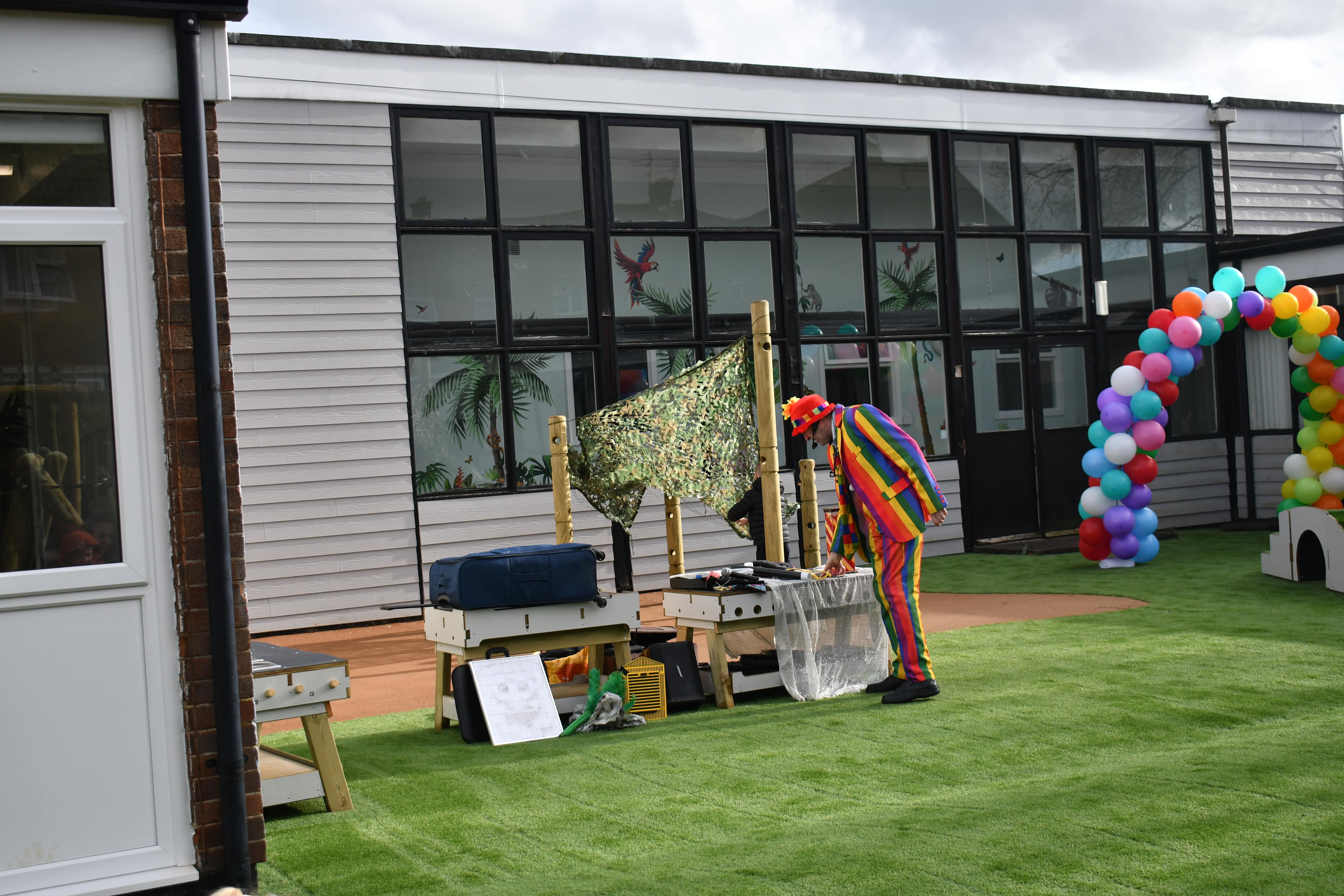 A man dressed in a rainbow suit and a red hat is looking at the science investigation area. A variety of toys are being set up by this man to encourage children to come investigate.