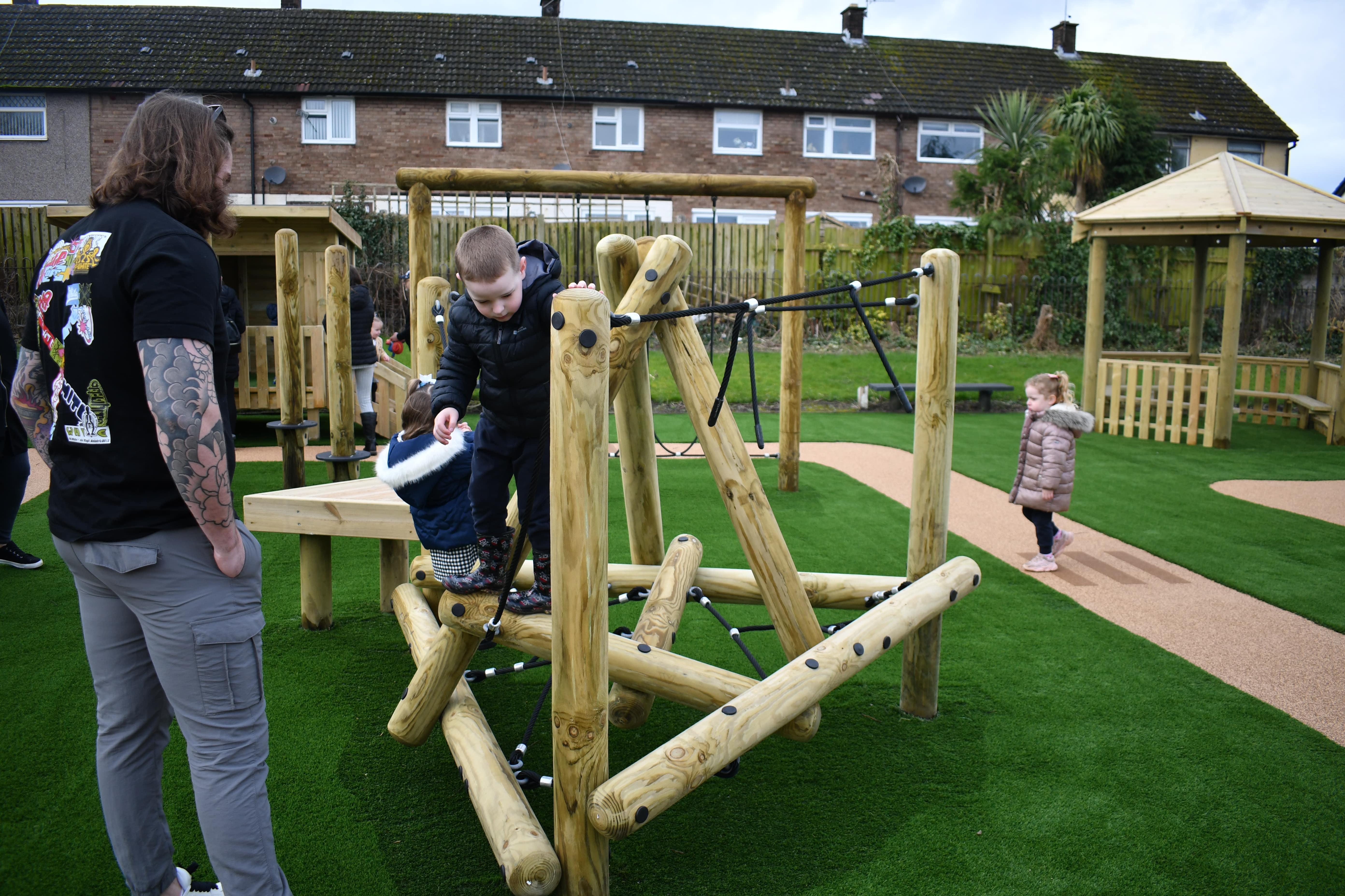 A Pinnacle Mount Climber has been installed on artificial grass, with a wetpour path to the left of the structure. A parent is watching their child climb on the commercial climbing frame.