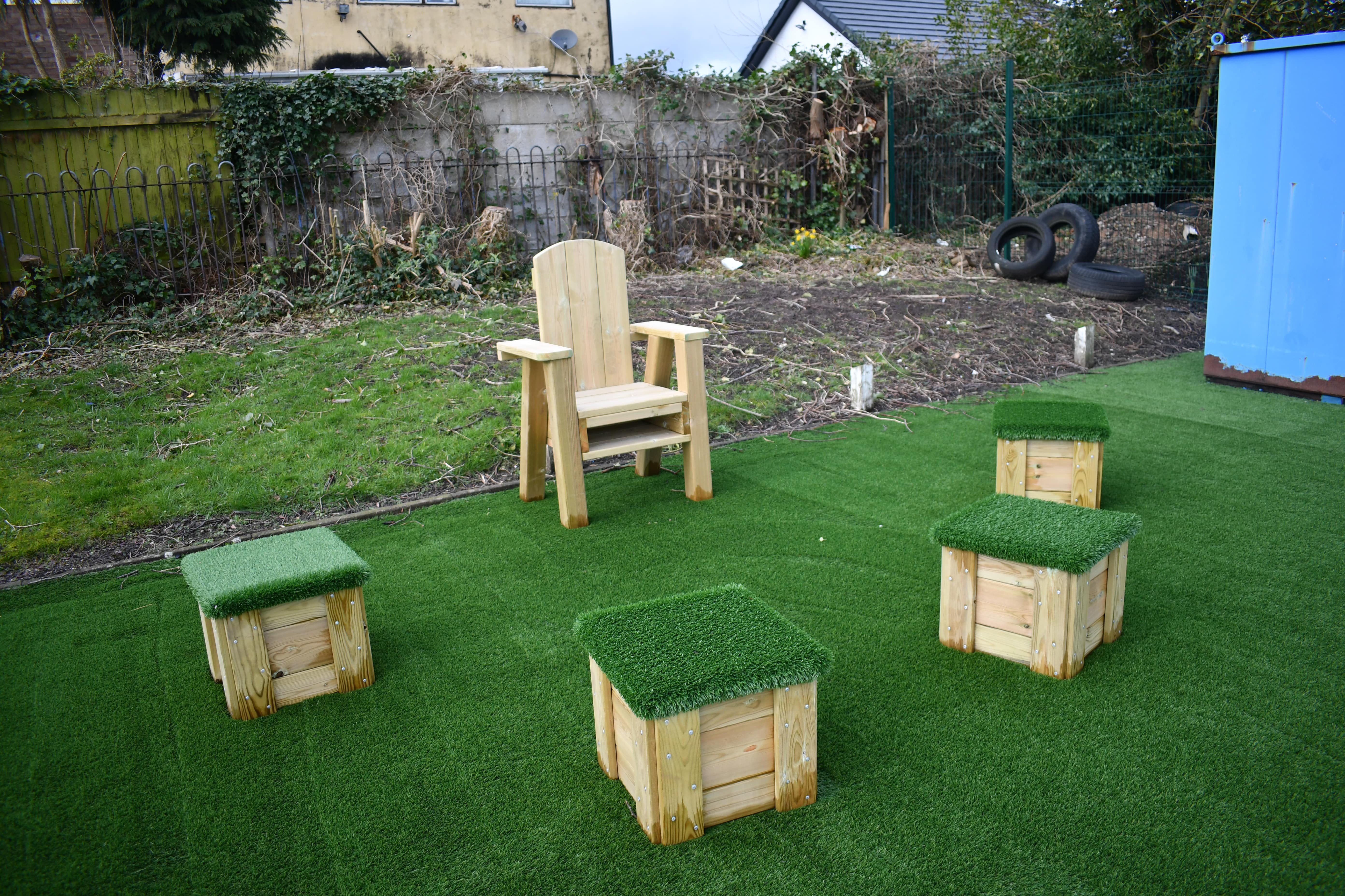 A freestanding wooden chair has been placed on the artificial grass surface, with 4 wooden blocks placed around the front of the chair. Each block has artificial grass placed on top so children can sit on them.